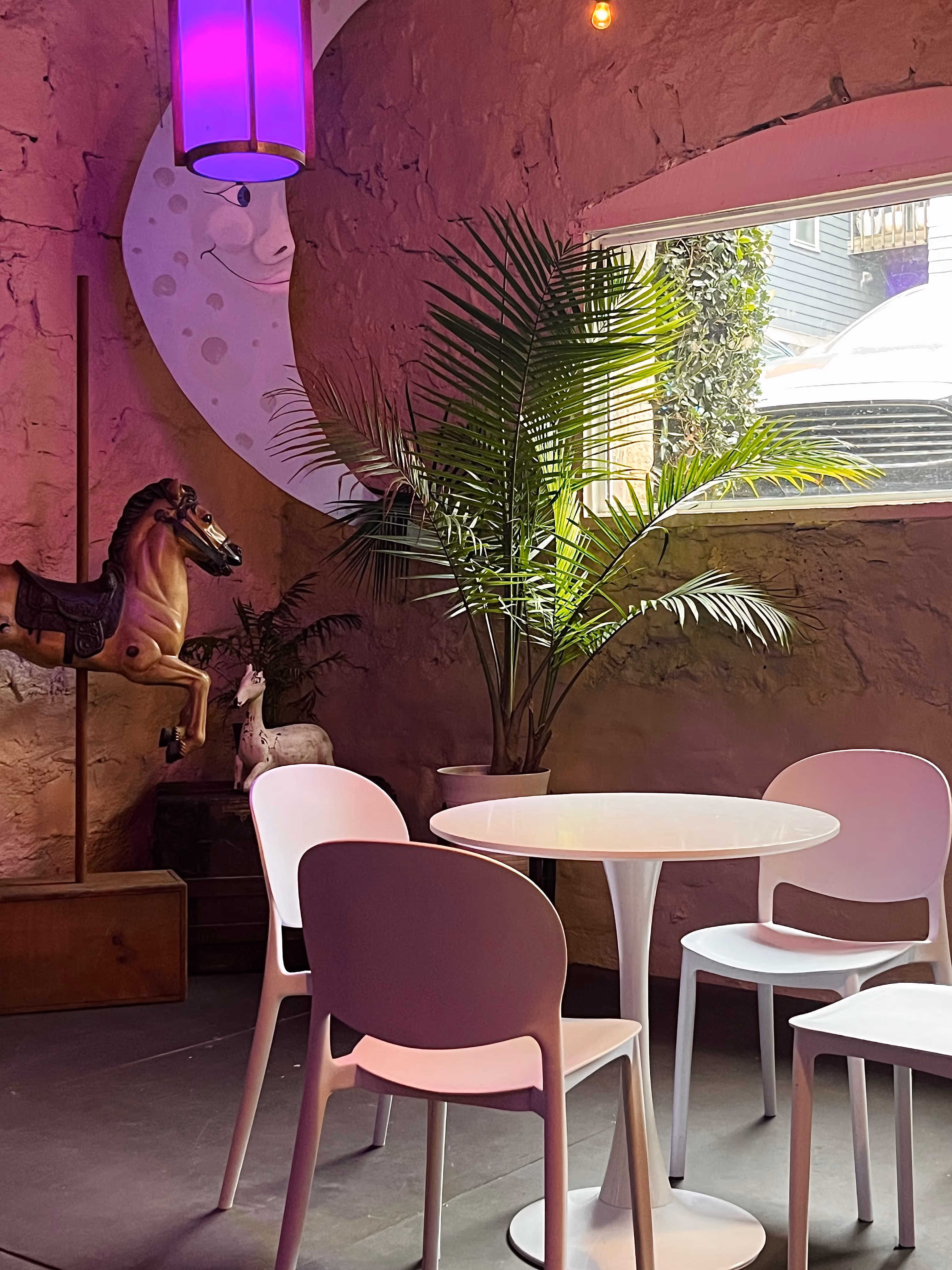 Event Space corner with white modern chairs and a round table beneath a purple hanging light, featuring a playful backdrop of a crescent moon mural, potted plants, and vintage carousel-style horse and deer figures.