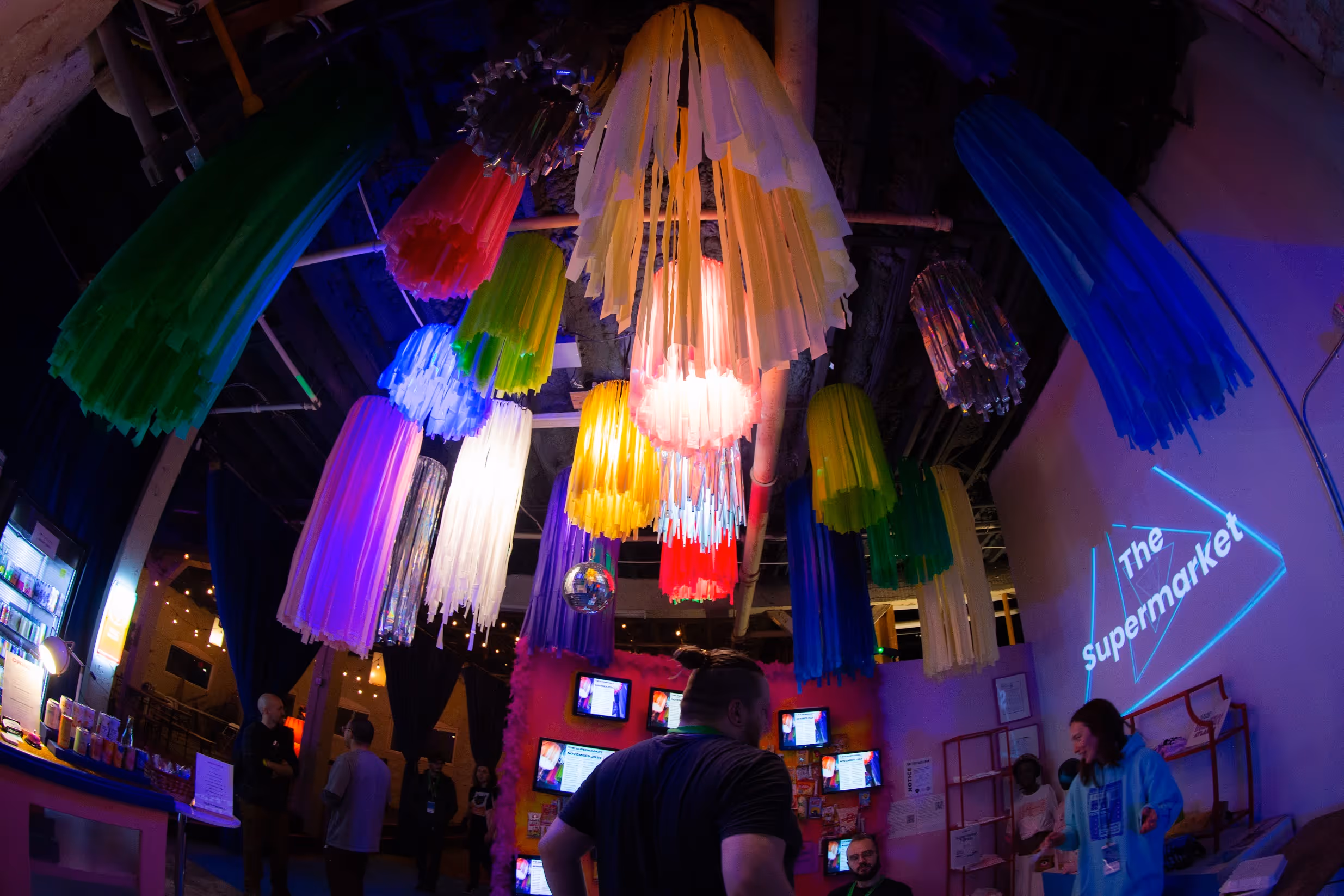 Ceiling view of an event space lit with colored hanging streamers and fabric strips in hues of green, blue, pink, and white, with a disco ball suspended among them.