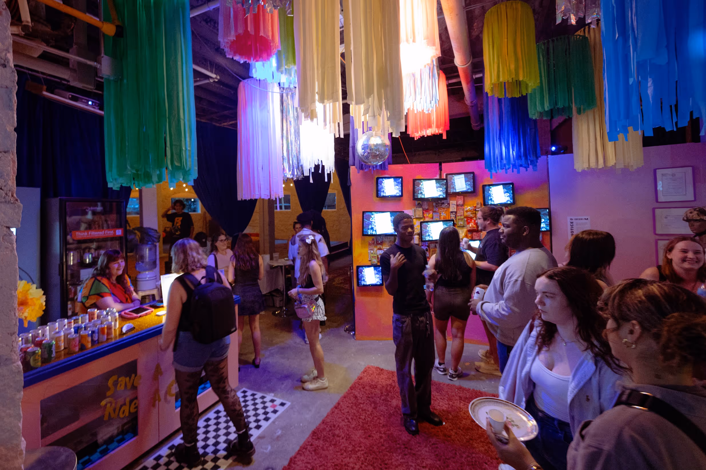 A colorful party or reception area with people mingling under vibrant hanging streamers, a disco ball overhead, and ambient lighting casting hues of pink, blue, and yellow throughout the space.