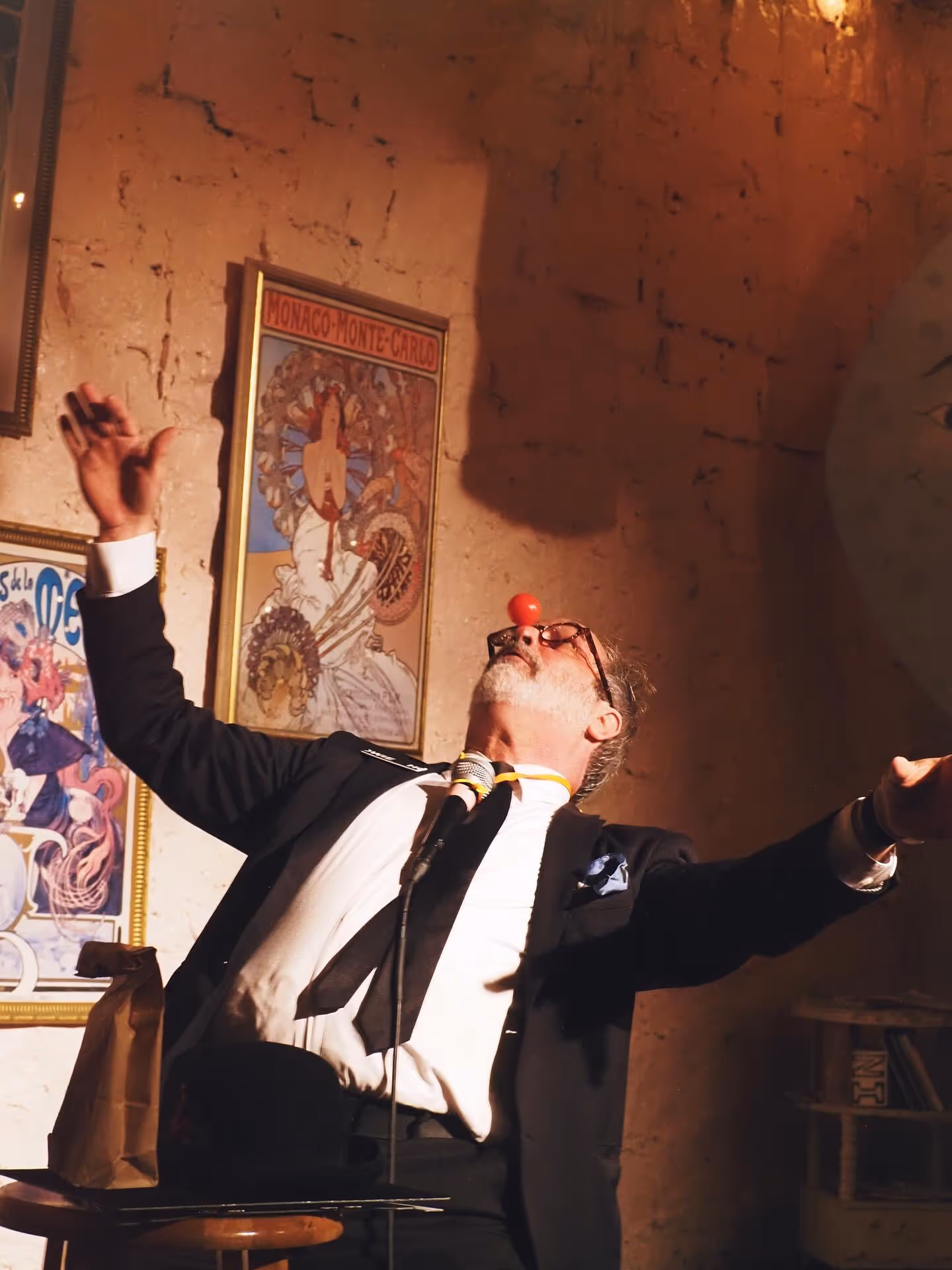 Performer in a suit on stage at The Supermarket, arms outstretched with a clown nose and microphone, mixing comedy and theatrical flair. Vintage posters hang on the wall behind. Secret Aisle blends magic, comedy, and absurdist performance in an intimate setting.
