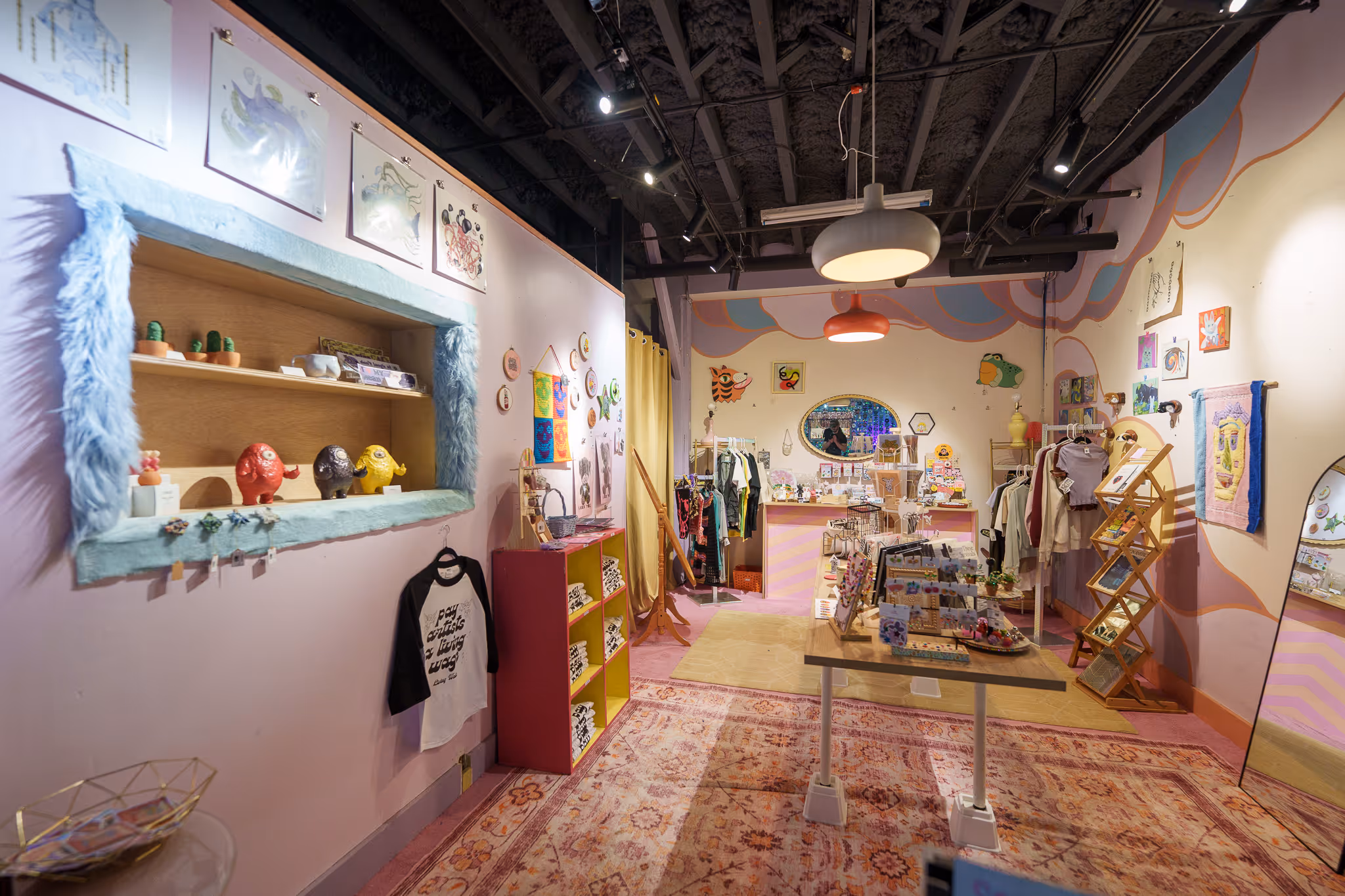 Colorful boutique store interior with pastel pink walls, eclectic clothing racks, decorative art pieces, and a patterned rug.