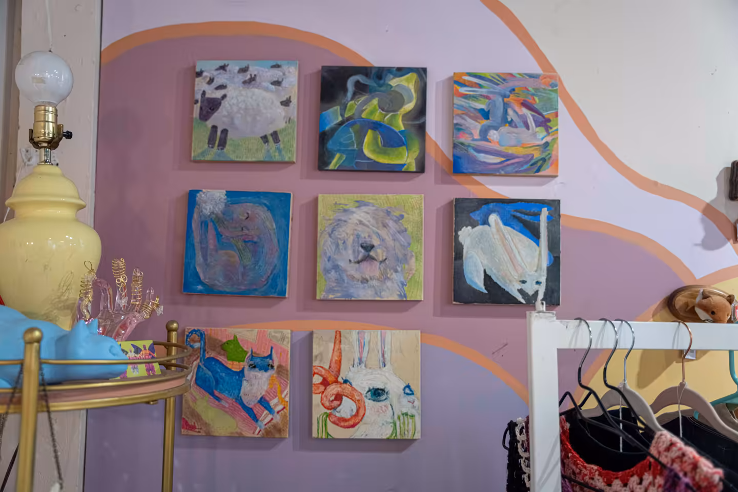Wall with nine colorful, abstract animal paintings arranged in three rows, featuring creatures like sheep, a lion, and other imaginative animals in a room with a pink and orange wavy mural background.