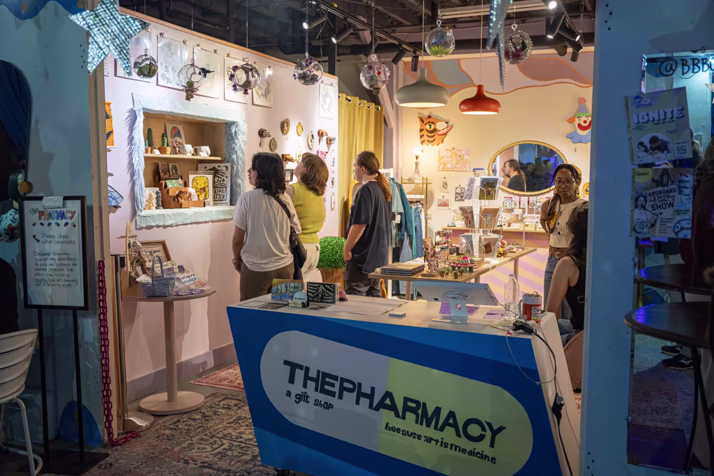 People browsing art and craft items inside The Pharmacy gift shop with colorful decor and hanging glass ornaments.