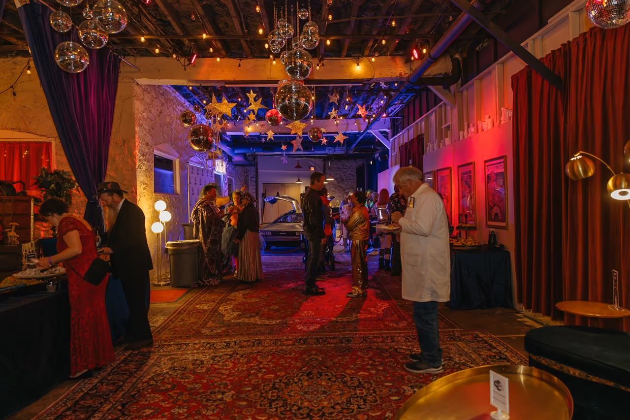 Indoor party with people mingling under hanging disco balls and star decorations with colorful lighting.