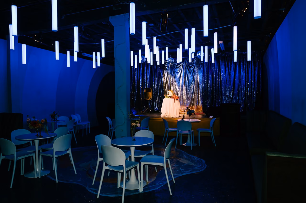 Dimly lit room with hanging tube lights, round tables with chairs, and a small stage with a draped table.
