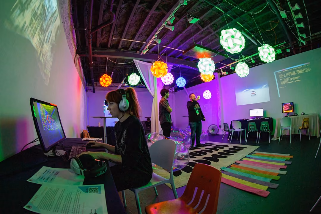 Person wearing headphones using a computer in a colorful, modern room with glowing hanging lights and others present.