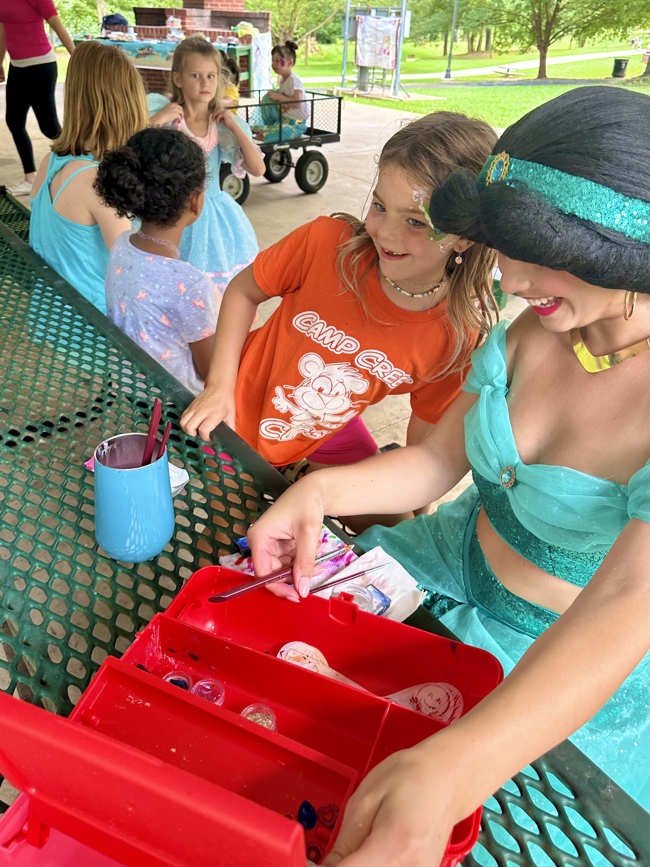 A young girl with face paint in an orange Camp Creek Cubs t-shirt smiles as a woman dressed as Princess Jasmine organizes face painting supplies on a green picnic table at an outdoor event.