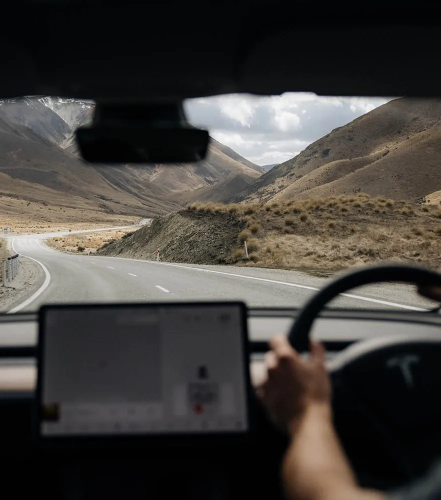 View from inside a Tesla driving on a winding road through dry, hilly terrain with mountains in the distance.
