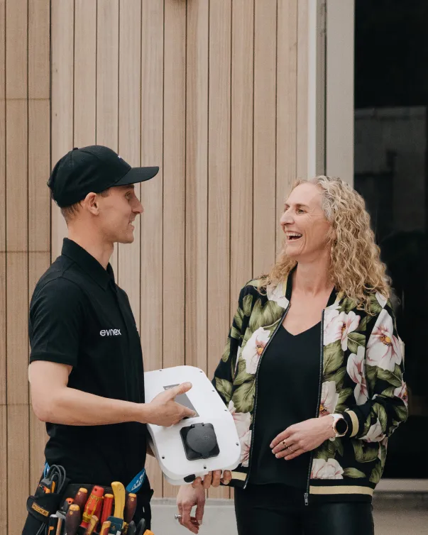 Electrician in black uniform holding an electrical device and talking with a smiling woman wearing a floral jacket.