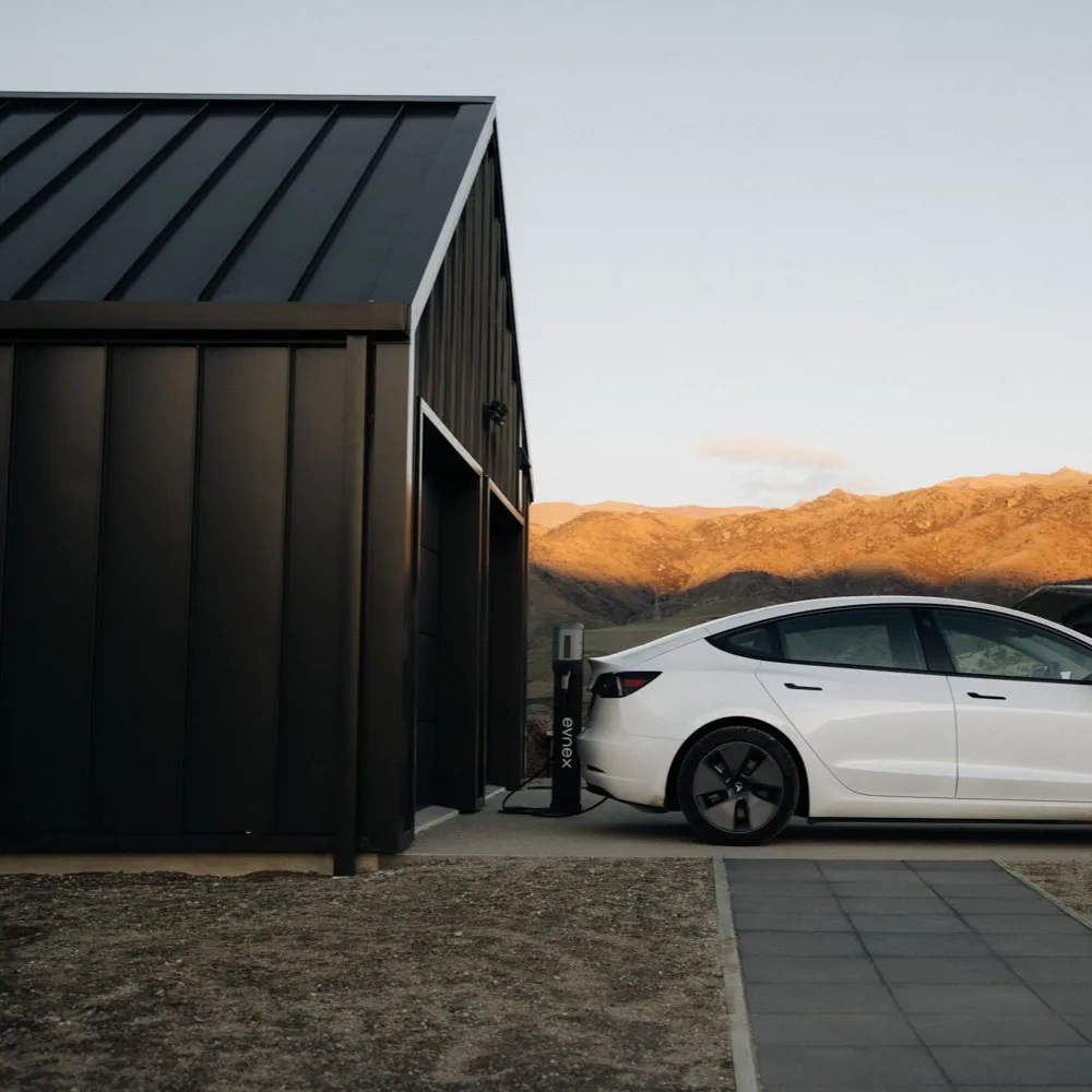 White electric car charging at an Evnex charging station next to a black modern building with hills in the background at sunset.