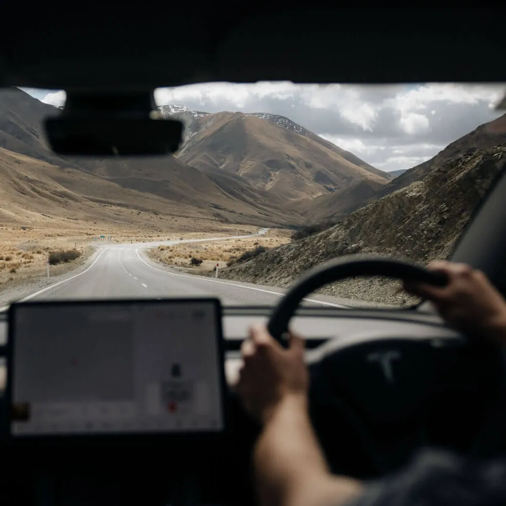 View from inside a Tesla car showing a person driving on a winding road through dry mountainous terrain under a cloudy sky.