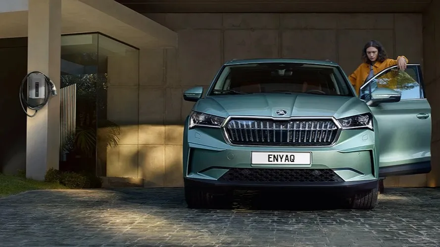 Front view of a green Škoda Enyaq electric car parked on a driveway with the driver's door open and a woman standing beside it.