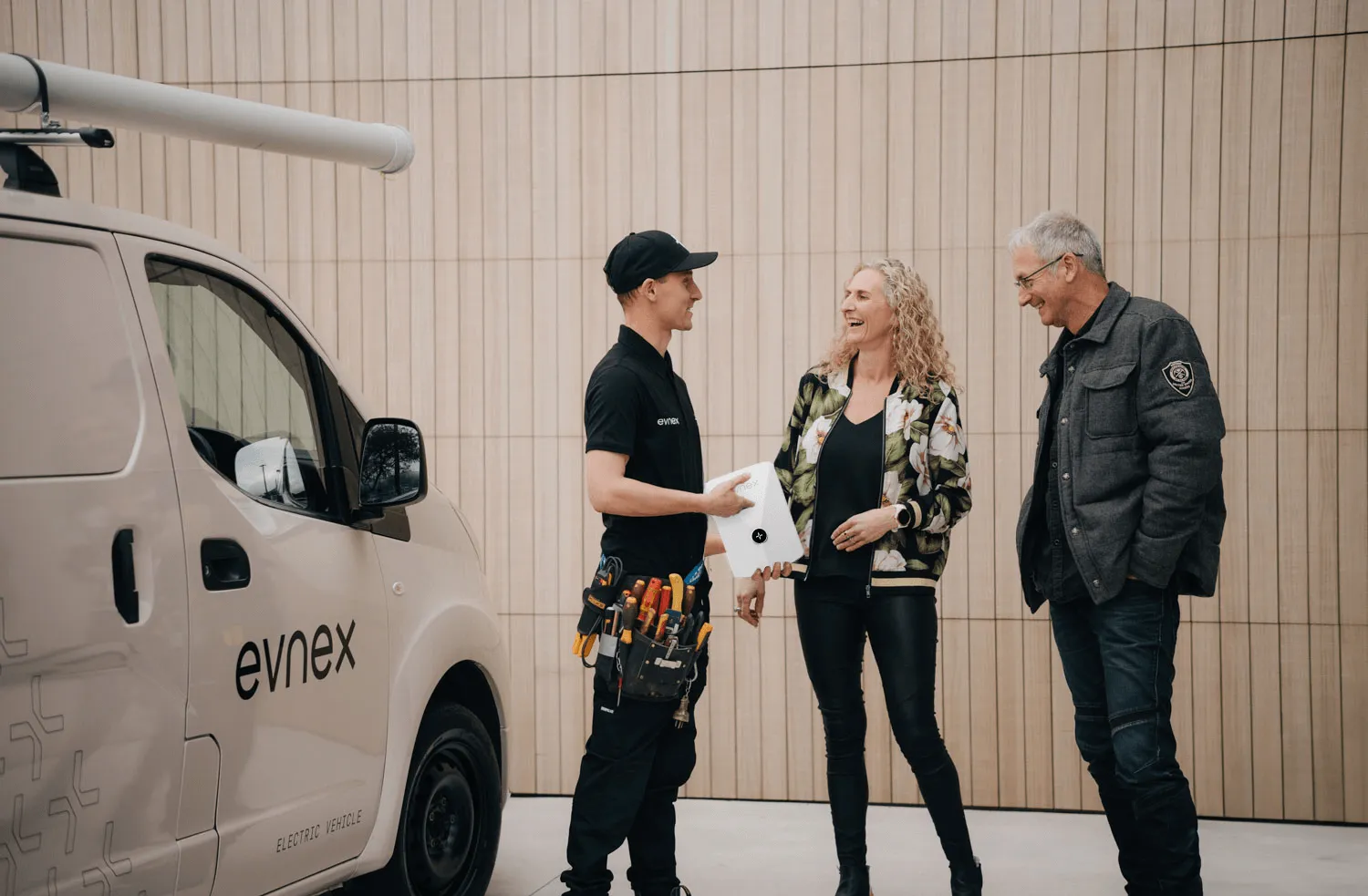 Technician in black uniform with tools talks to a smiling couple beside an Evnex electric vehicle.