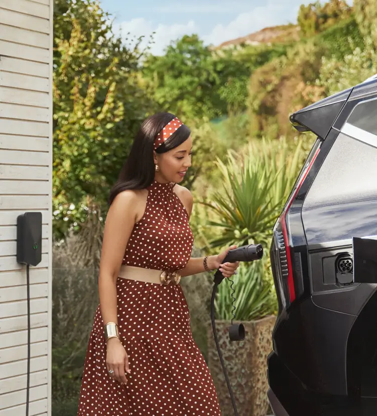 Woman in a brown polka dot dress plugging an electric car charger into a vehicle outside near greenery.