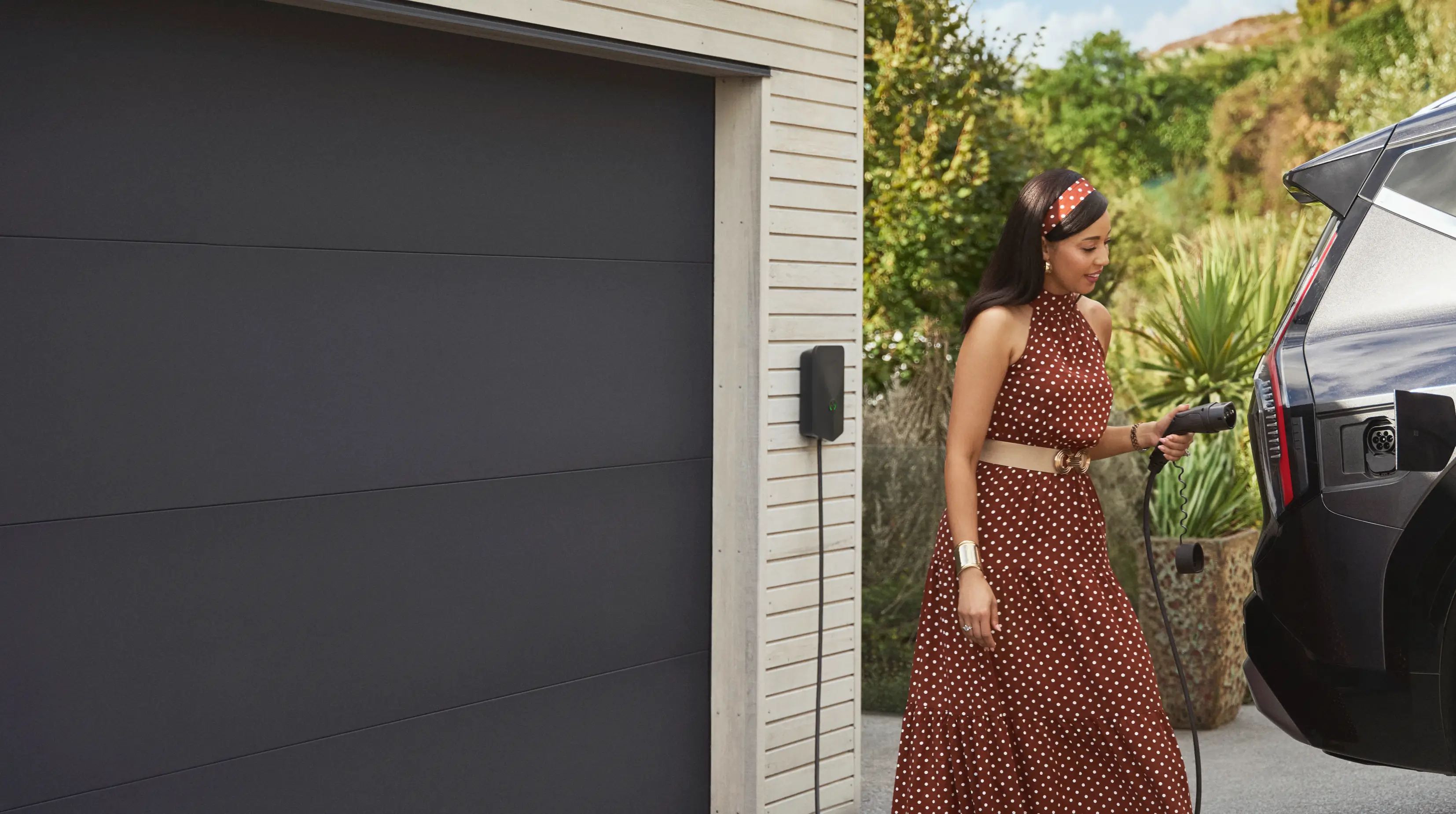 Woman in a brown polka dot dress plugging a cord into a black electric vehicle parked near a house garage.
