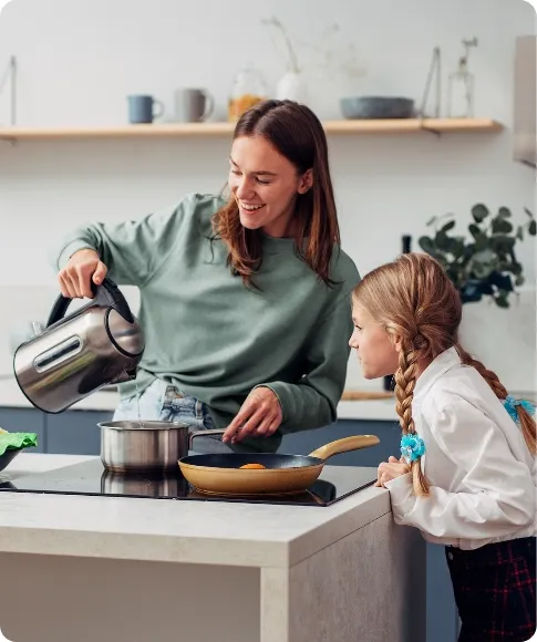 A woman pours water from a kettle into a pot while a young girl with braided hair watches in a modern kitchen.