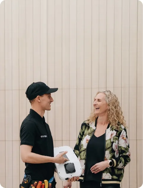 An electrician in a black uniform is speaking with a smiling woman, while holding an electrical device.