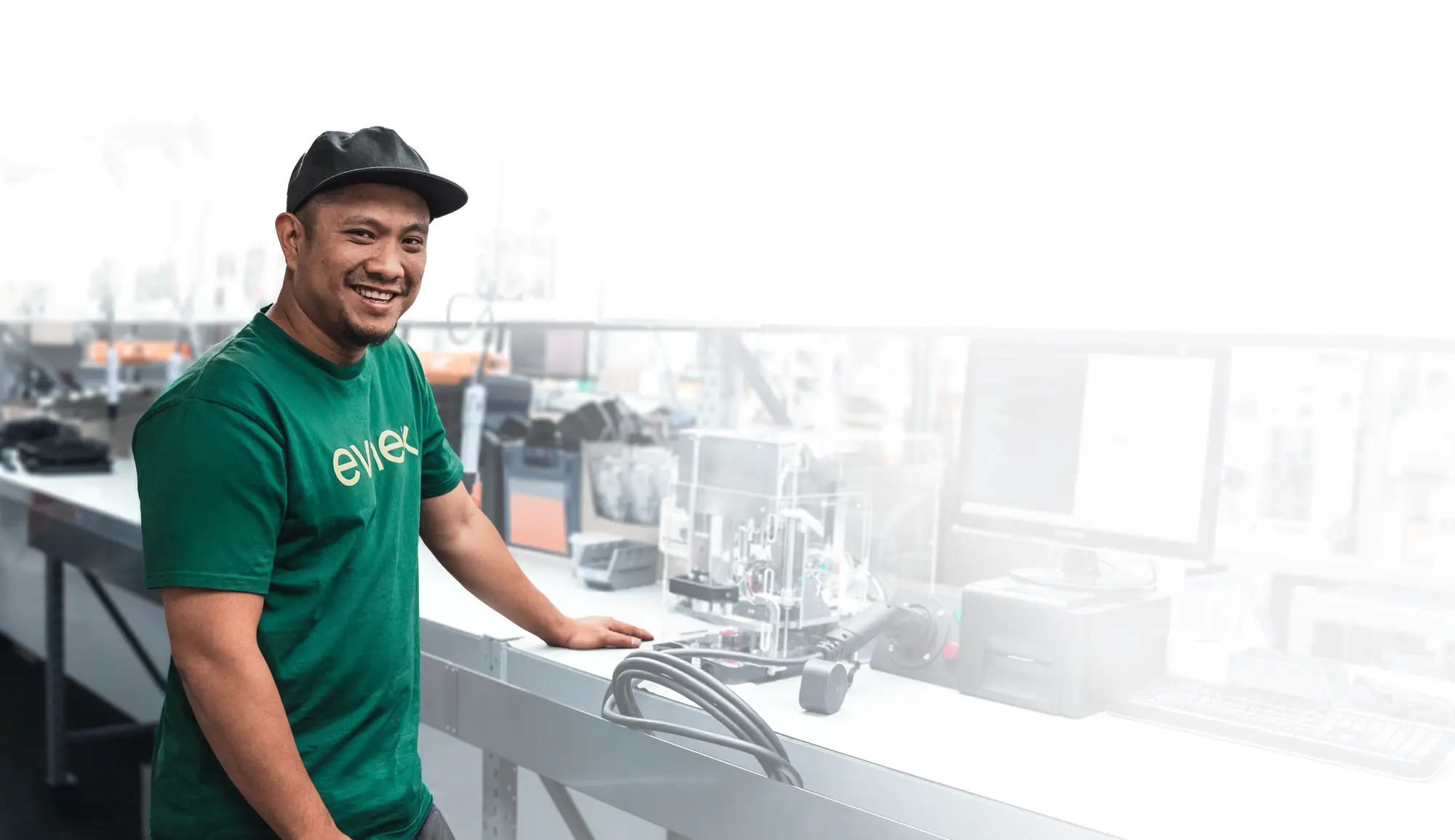 A smiling man wearing a green Evnex t-shirt and a black cap stands next to a workbench with electronic equipment and a computer monitor.