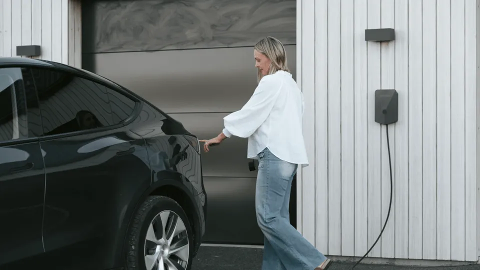 A blonde Pākehā woman in light blue, baggy jeans and a long-sleeved white shirt plugs in her black EV sedan. She is using a Volcanic-coloured Evnex charger set against white weatherboard on the exterior of the garage.