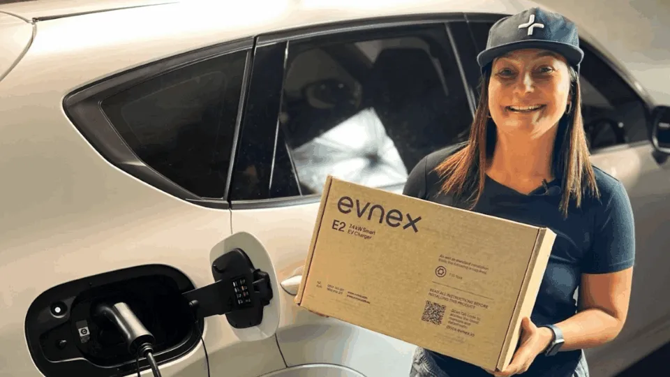 Cintia, a smiling woman with brown hair, wears a black Evnex cap and a green Evnex shirt. She is standing in front of a white plugged-in EV, holding the recycled cardboard box her charger came in.