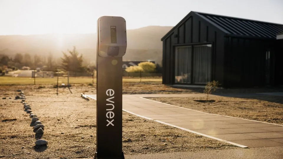 In the background, a black barn-like house and a wide open tussock with a low sun illuminating the ground and treeline in the distance. In the foreground, an Evnexx X22 sits on a black pedestal, backlit by the sinking sun.
