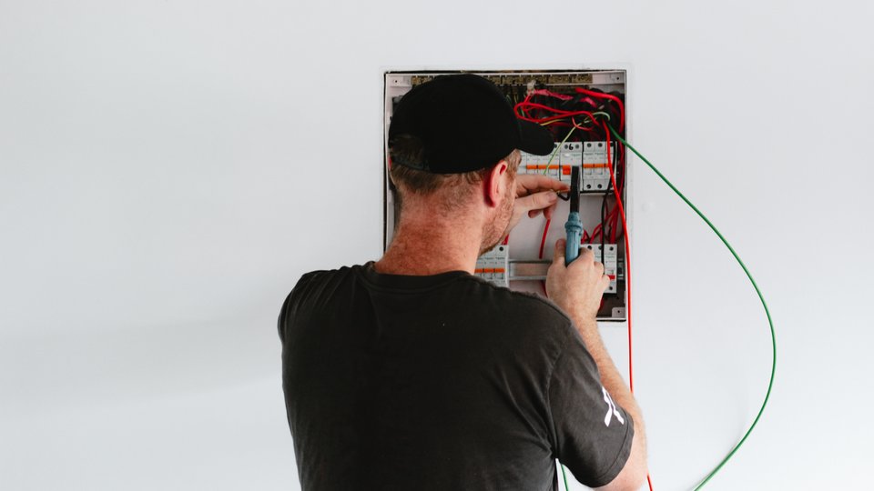 The back top half of an electrician (a ginger man wearing a black t-shirt and black baseball cap) is in the foreground. he's fiddling with a switchboard, which is set inside a blank white wall.