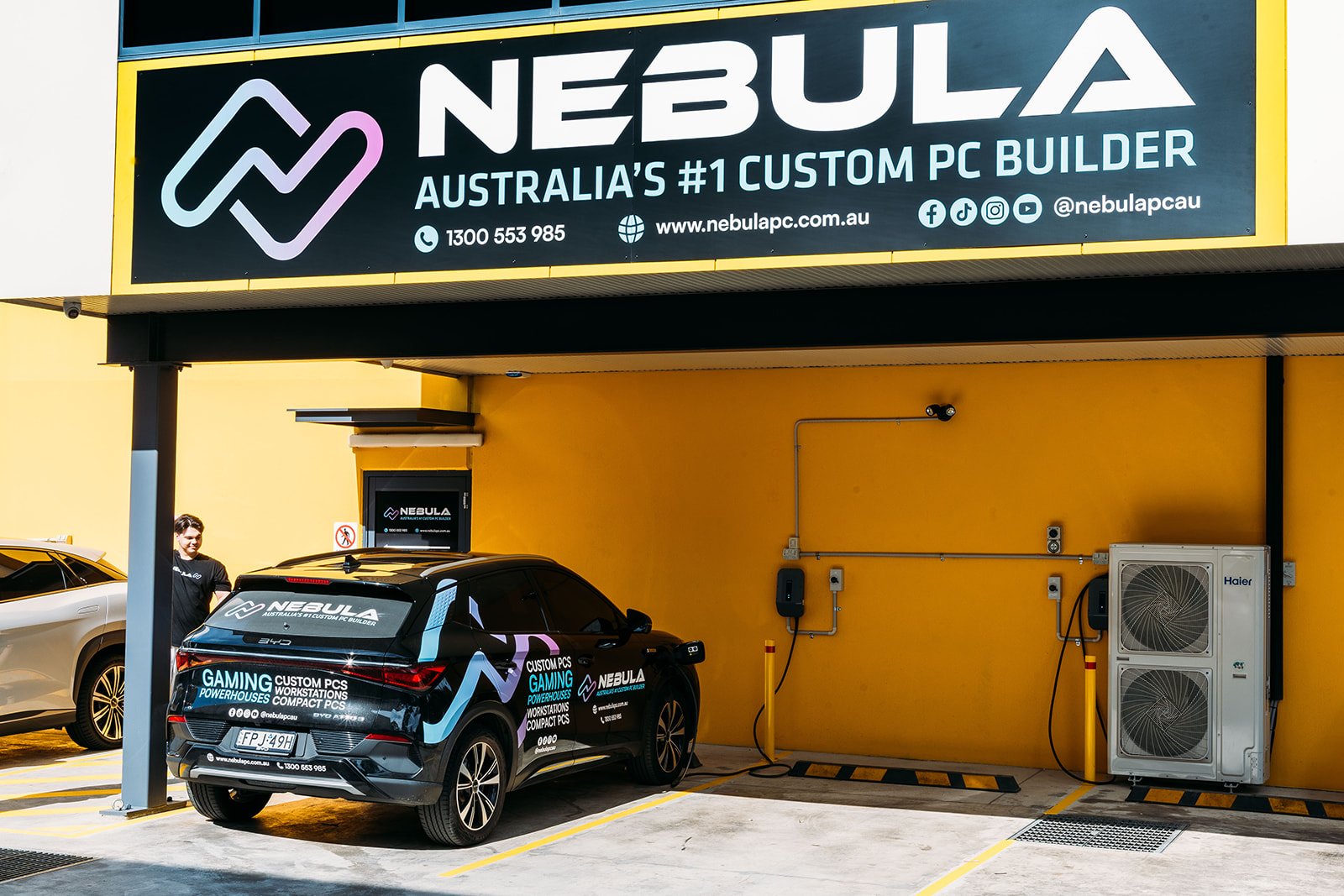 A black BYD is parked outside Nebula PC's bright yellow front wall. Two black X22s are attached the front of the building. The sun is shining brightly down on the car (on the left of the image) and the empty parks to the right of it.
