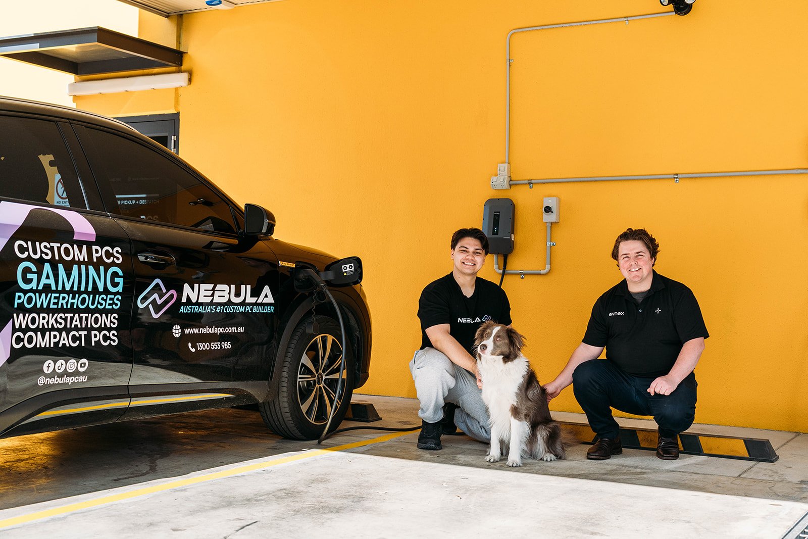 Crouched and smiling on the ground next to a plugged-in black BYD are, from left to right: Ryan from Nebula PC, Steve Nebula PC's resident dog, and Mitch Arnold from Evnex.i
