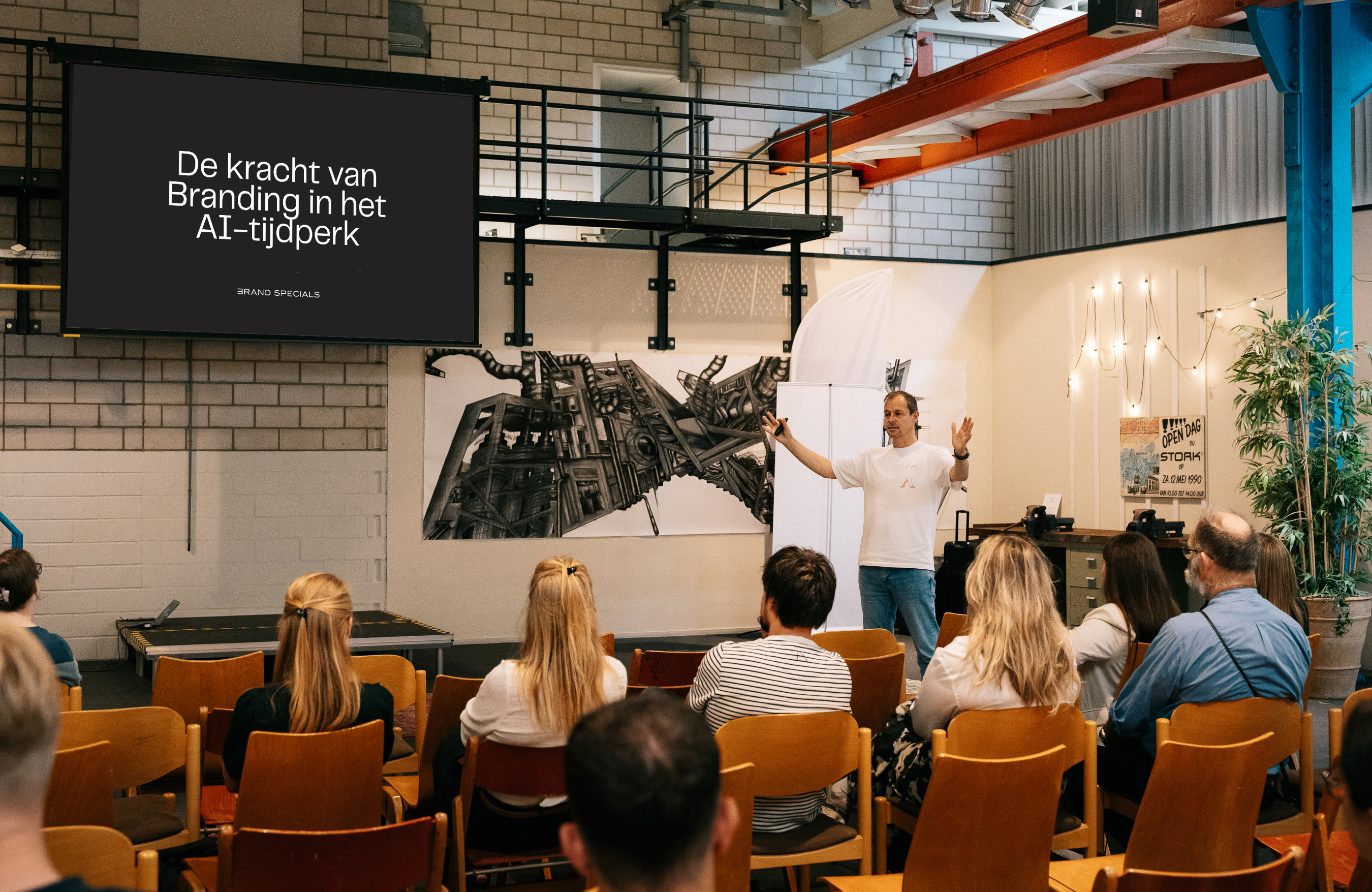 Man in a white t-shirt giving a presentation to an audience seated in wooden chairs in an industrial-style room with brick walls, a large screen displaying Dutch text about branding in the AI era, and artwork on the wall behind him.
