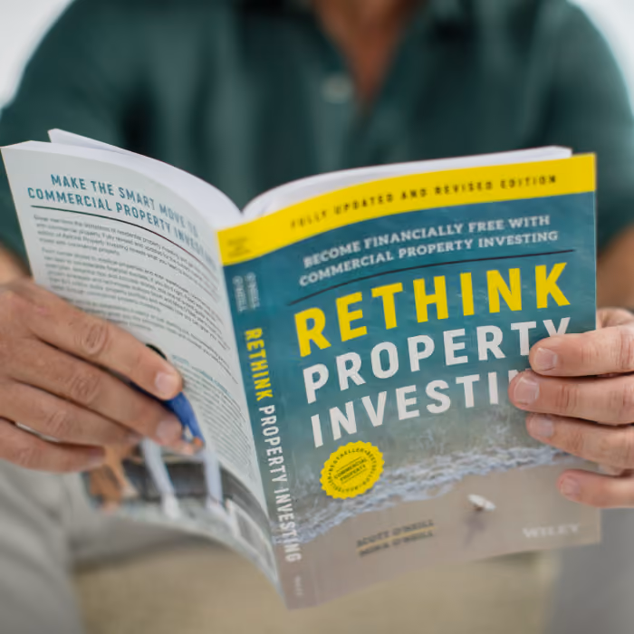 Person holding and reading a book titled 'Rethink Property Investing' with a cover showing a beach scene and the subtitle 'Become financially free with commercial property investing.'