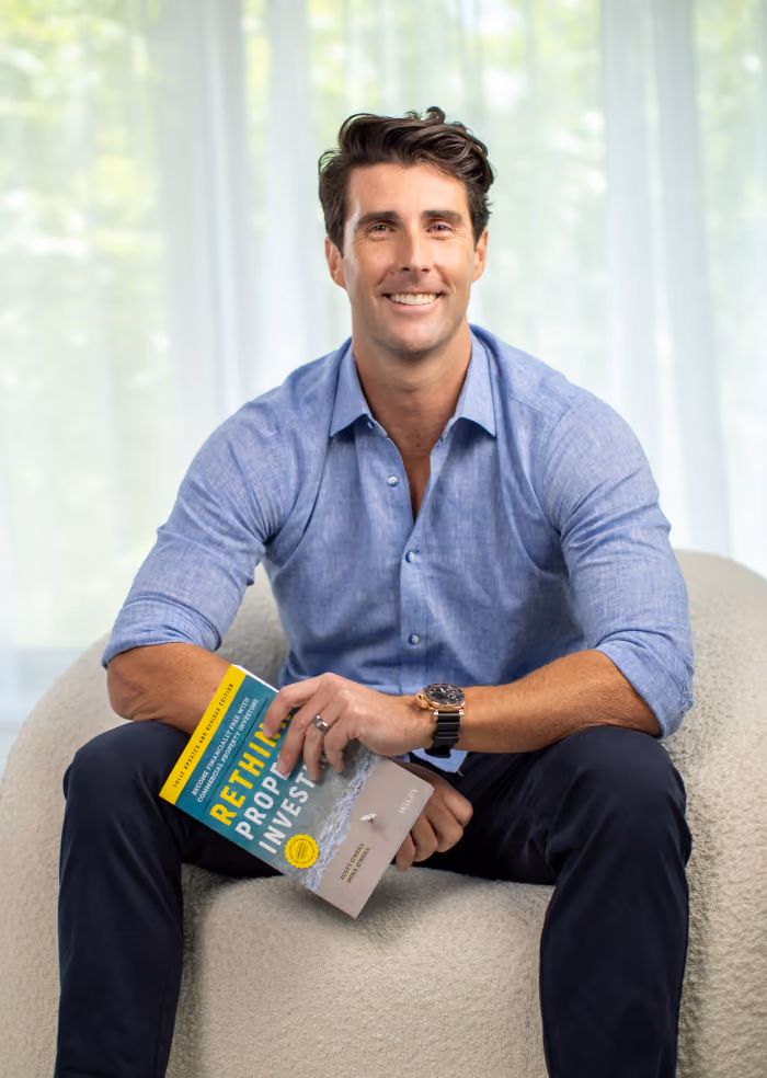 Smiling man with dark hair wearing a blue button-down shirt and dark pants, seated on a light-colored textured chair holding a book titled 'RETHINK PROP INVEST'.