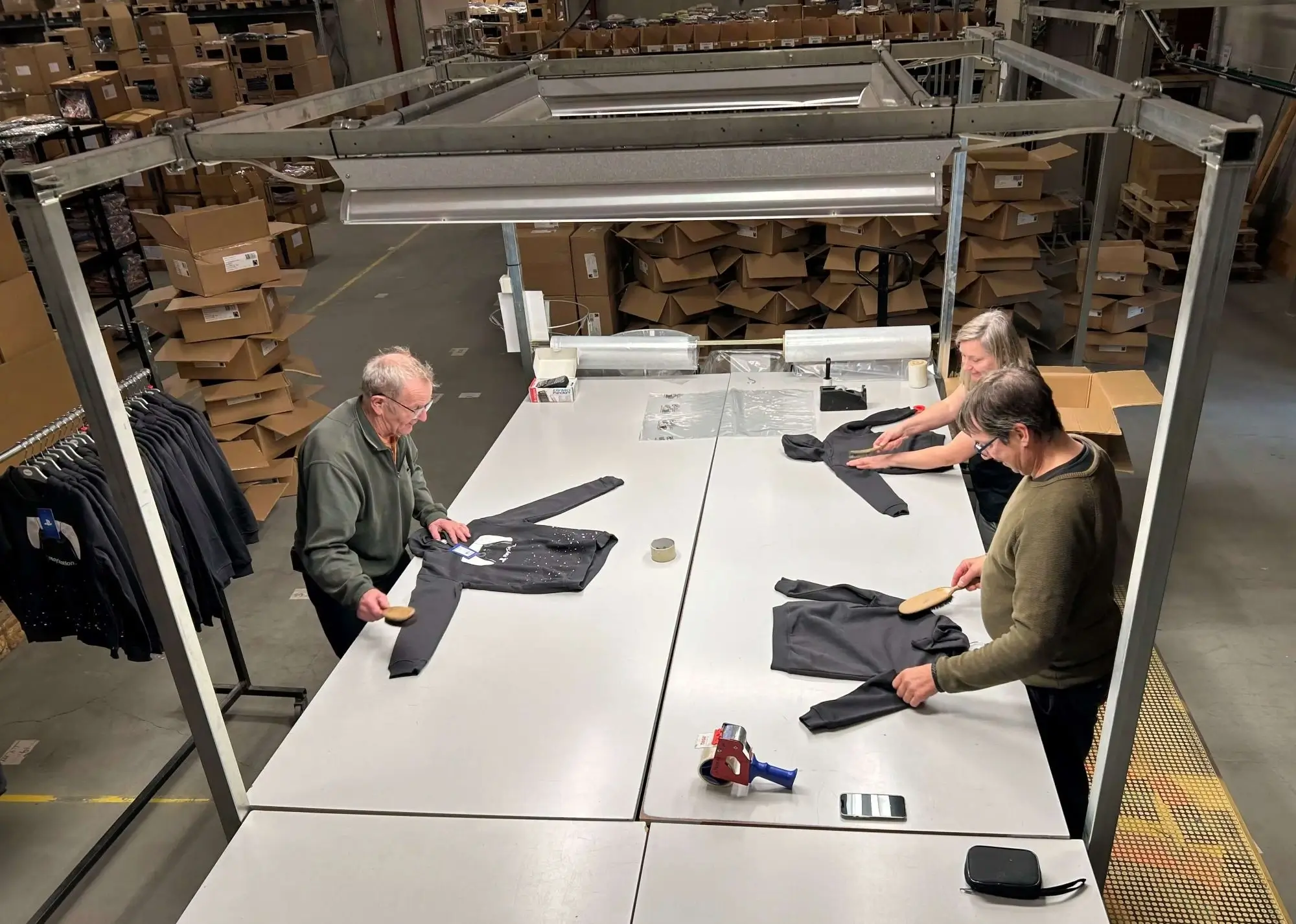 Three people brushing black sweatshirts at a large white table in a warehouse filled with cardboard boxes.
