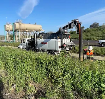 A wight parked truck of Pacific West Construction on the side of the road, with workers in hard hats and machinery in the background.