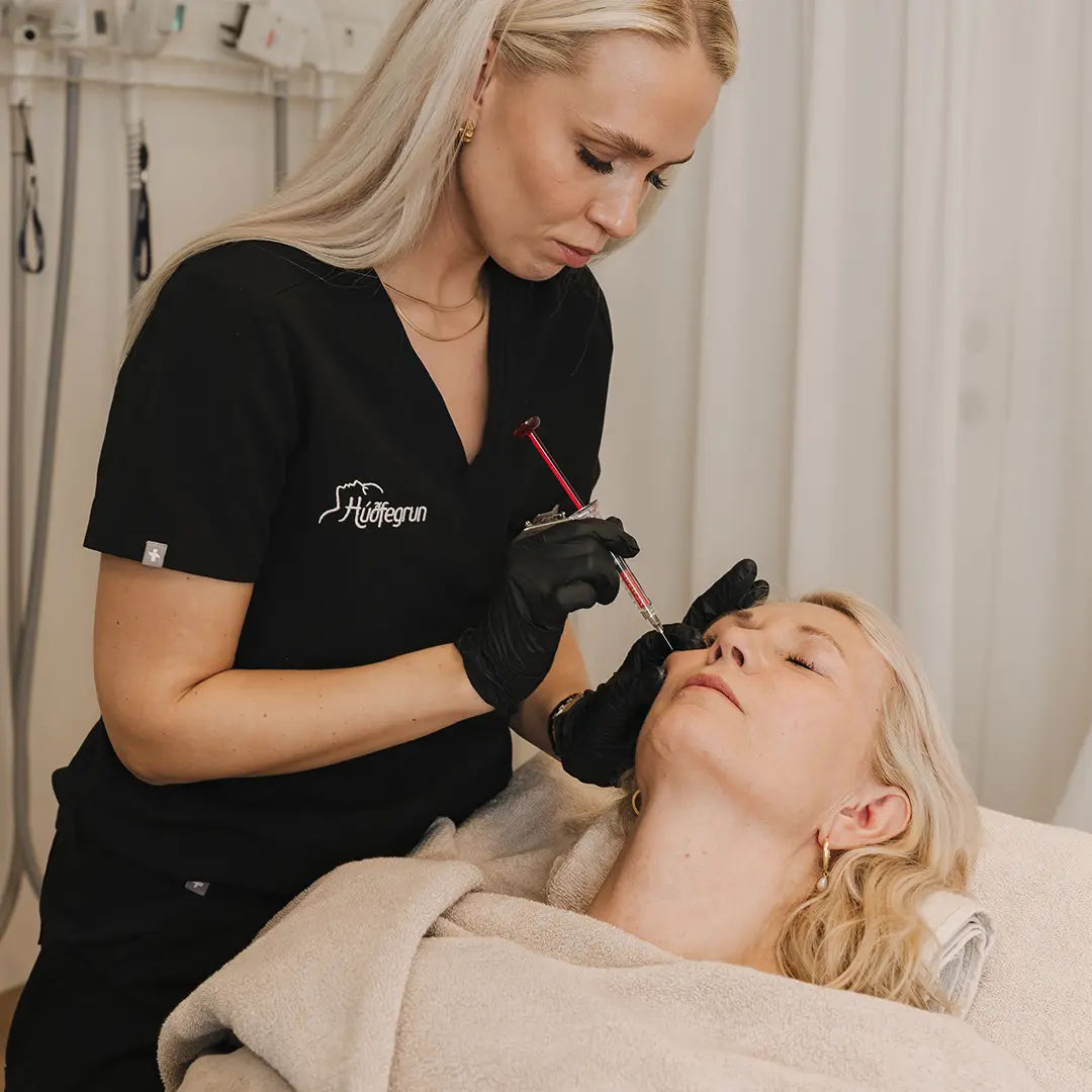 Hjúkrunarfræðingur framkvæmir kinnfyllingu með hýalúrónsýru-fylliefni sem brotnar niður á náttúrulegan hátt í líkamanum. Skjólstæðingur liggur afslappaður með hlutlausan svip á meðan meðferðin fer fram, og í bakgrunni má sjá hluta af háþróaðri lækninga- og lasertækni Hudfegrun.