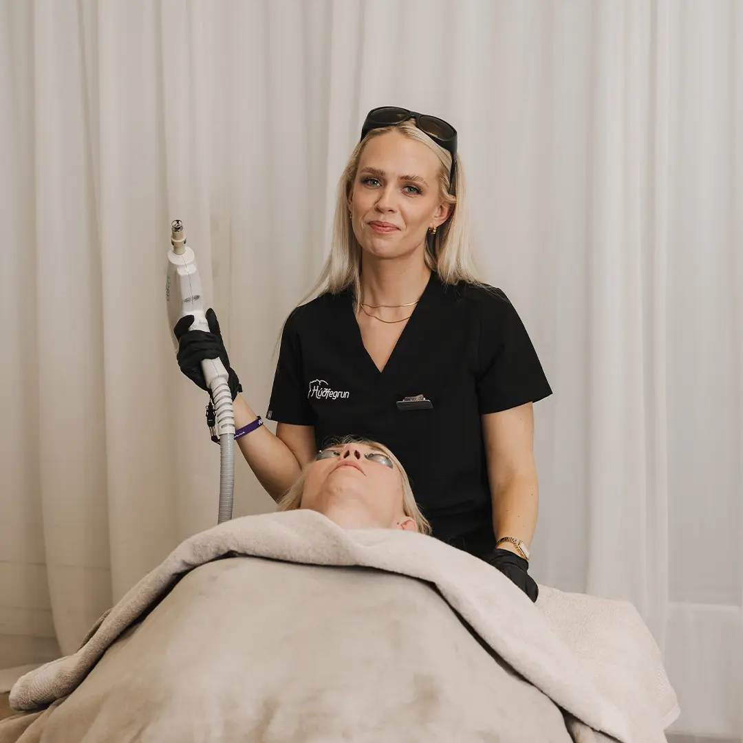 Hjúkrunarfræðingur hjá Hudfegrun heldur á leysitæki fyrir laserlyftingu á andliti og hálsi á meðan skjólstæðingur liggur tilbúinn með augnhlífar. Myndin er tekin í björtum og hreinum aðstæðum kliníkurinnar þar sem öryggi og fagmennska eru í forgrunni.