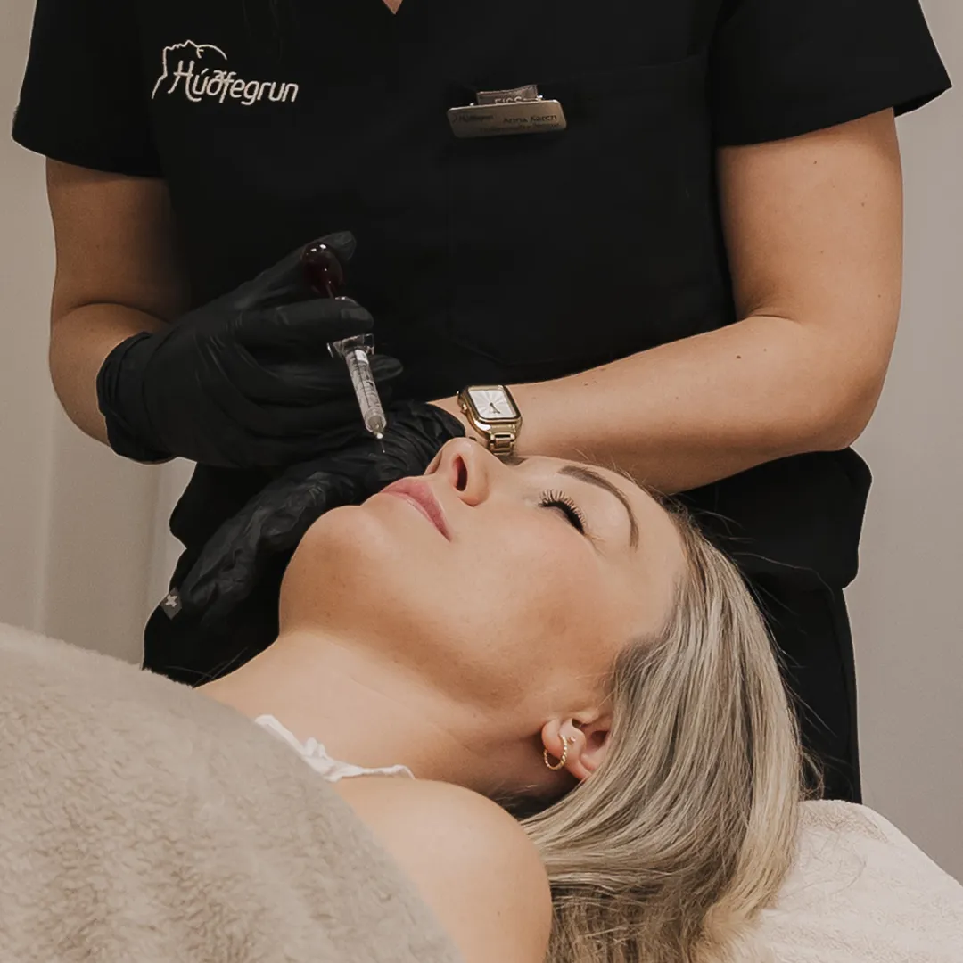 Sérfræðingur hjá Húðfegrun framkvæmir Toxín meðferð á andliti konu með nákvæmri sprautun til að draga úr fínum línum og hrukkum. Meðferðin slakar á vöðvum í andliti og gefur sléttari, ferskari og unglegri húð með sýnilegum árangri eftir nokkra daga.