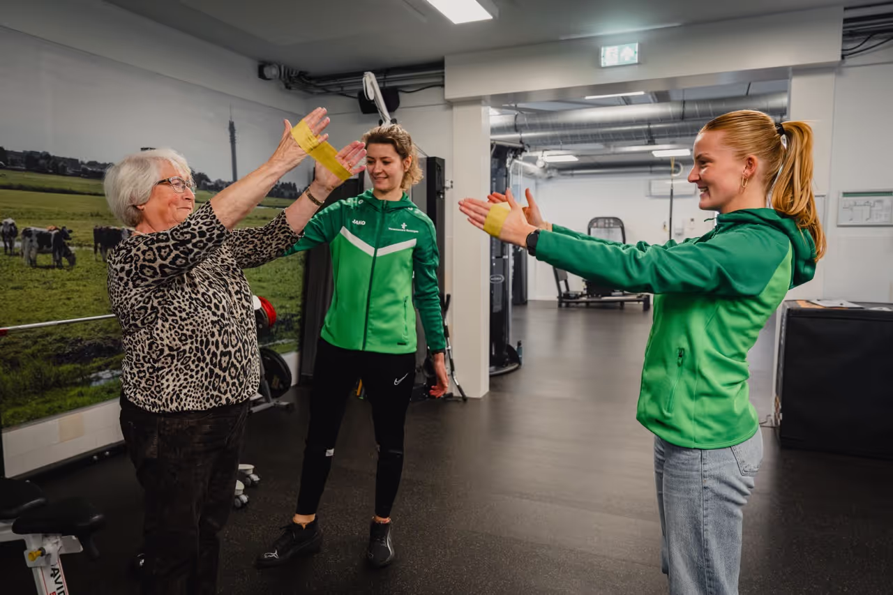 Two women in green jackets guiding an elderly woman doing arm exercises with yellow resistance bands in a gym.