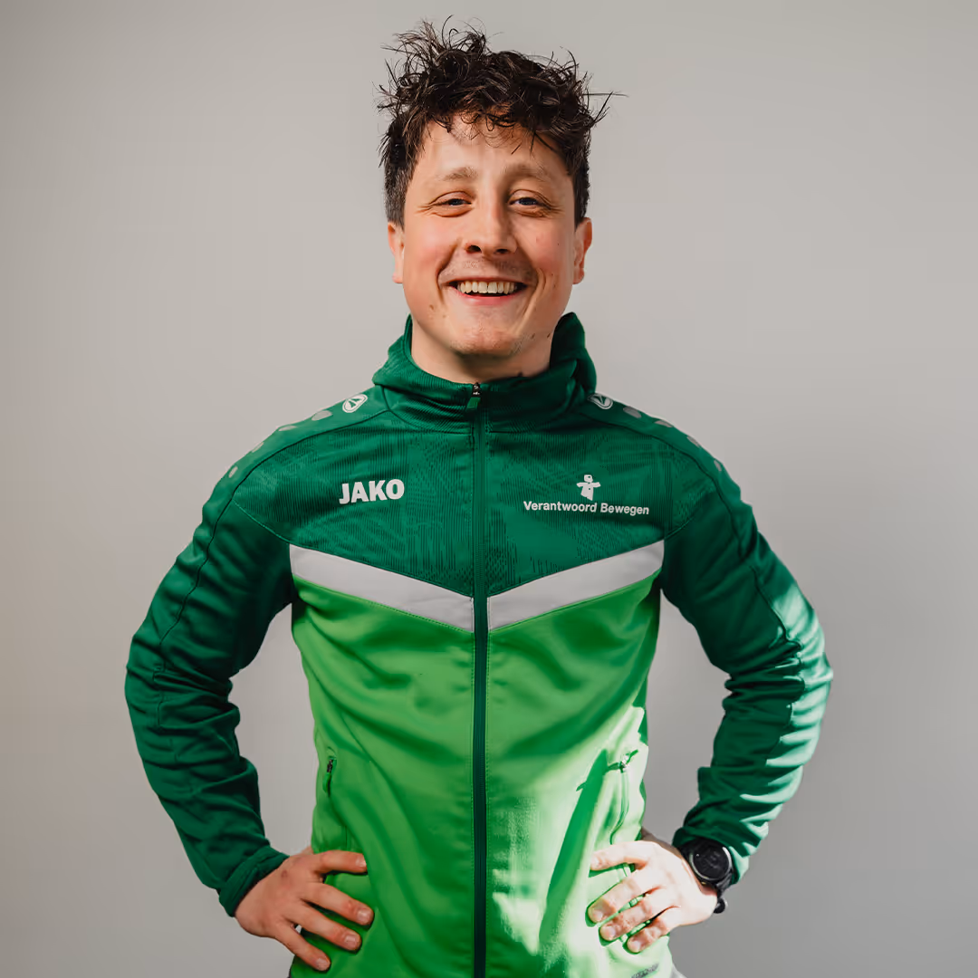 Smiling man with curly hair wearing a green and dark green sports jacket with hands on hips against a plain background.