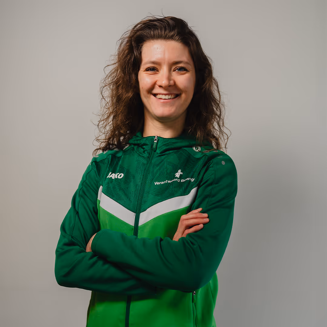 Smiling woman with curly brown hair wearing a green sports jacket with arms crossed against a plain gray background.