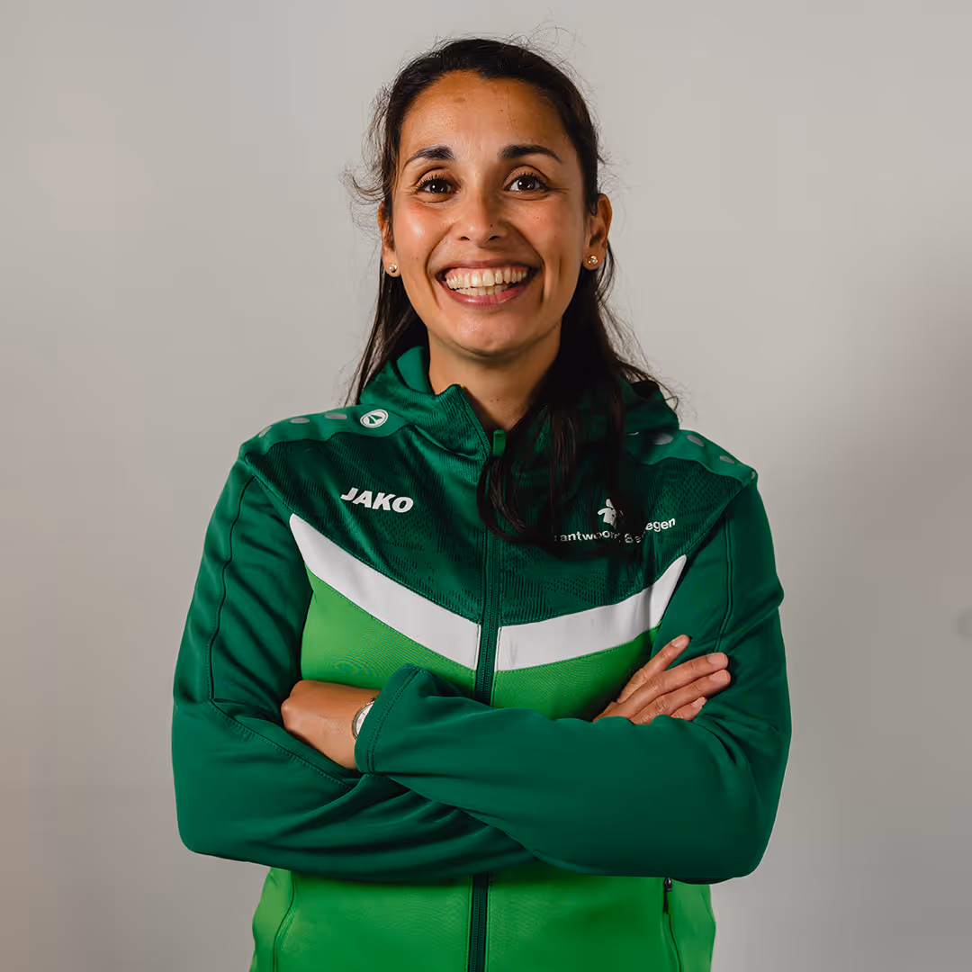 Smiling woman with long dark hair wearing a green sports jacket with crossed arms against a plain light background.