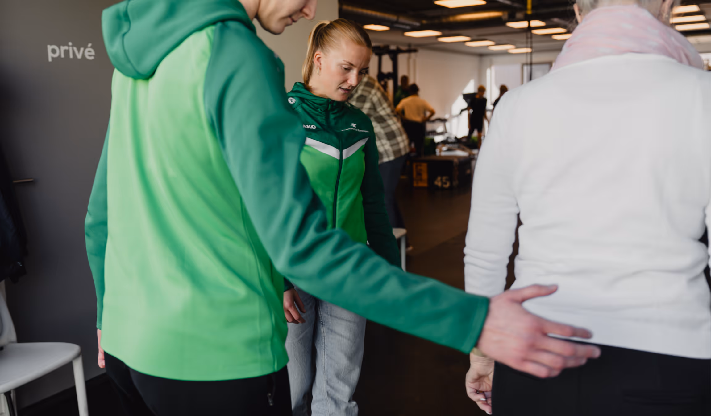 A person in a green jacket guides an older person in a white sweater in a gym setting with other people and exercise equipment in the background.