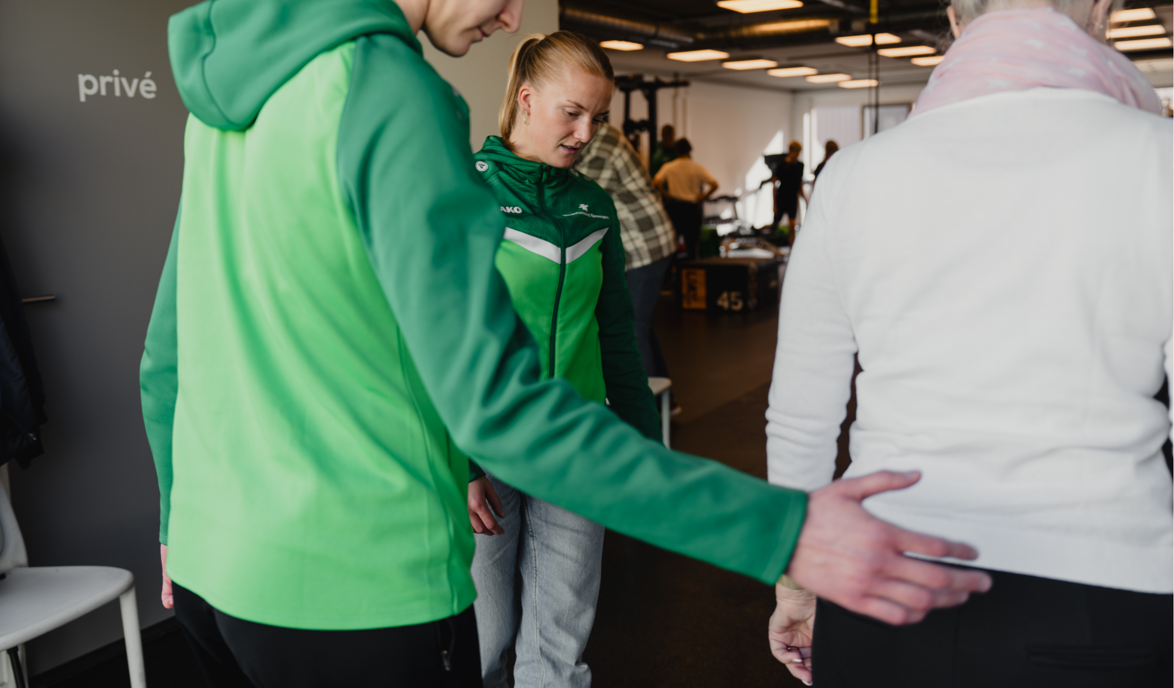 A person in a green jacket guides an older person in a white sweater in a gym setting with other people and exercise equipment in the background.