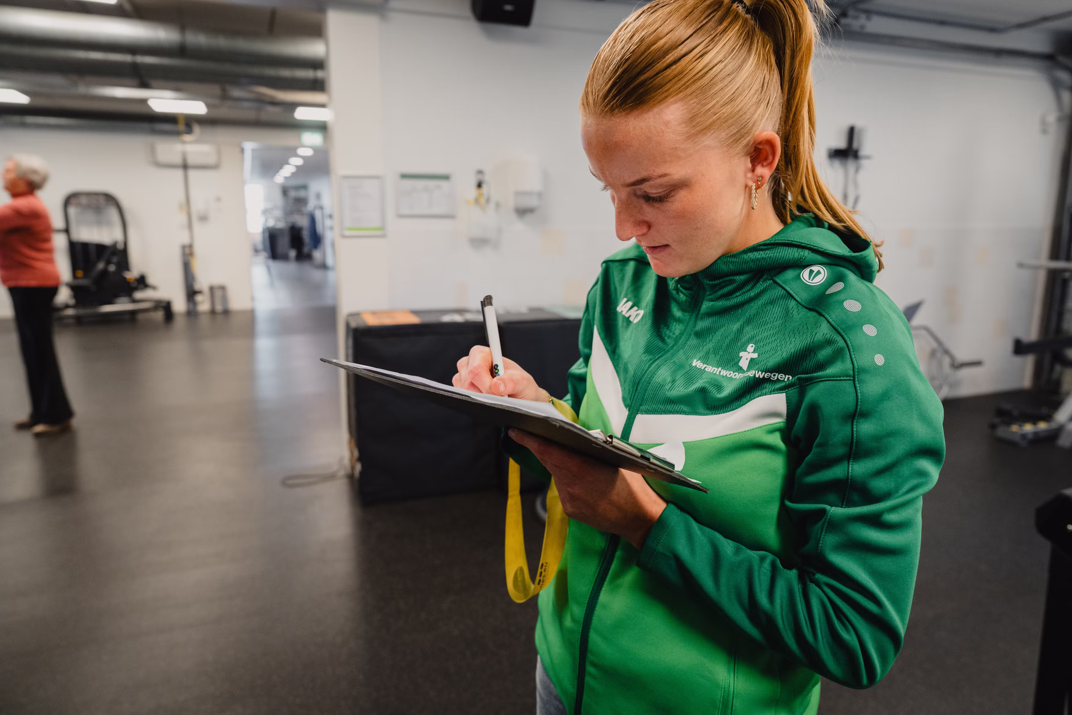 Young woman in a green jacket writing on a clipboard inside a gym setting.