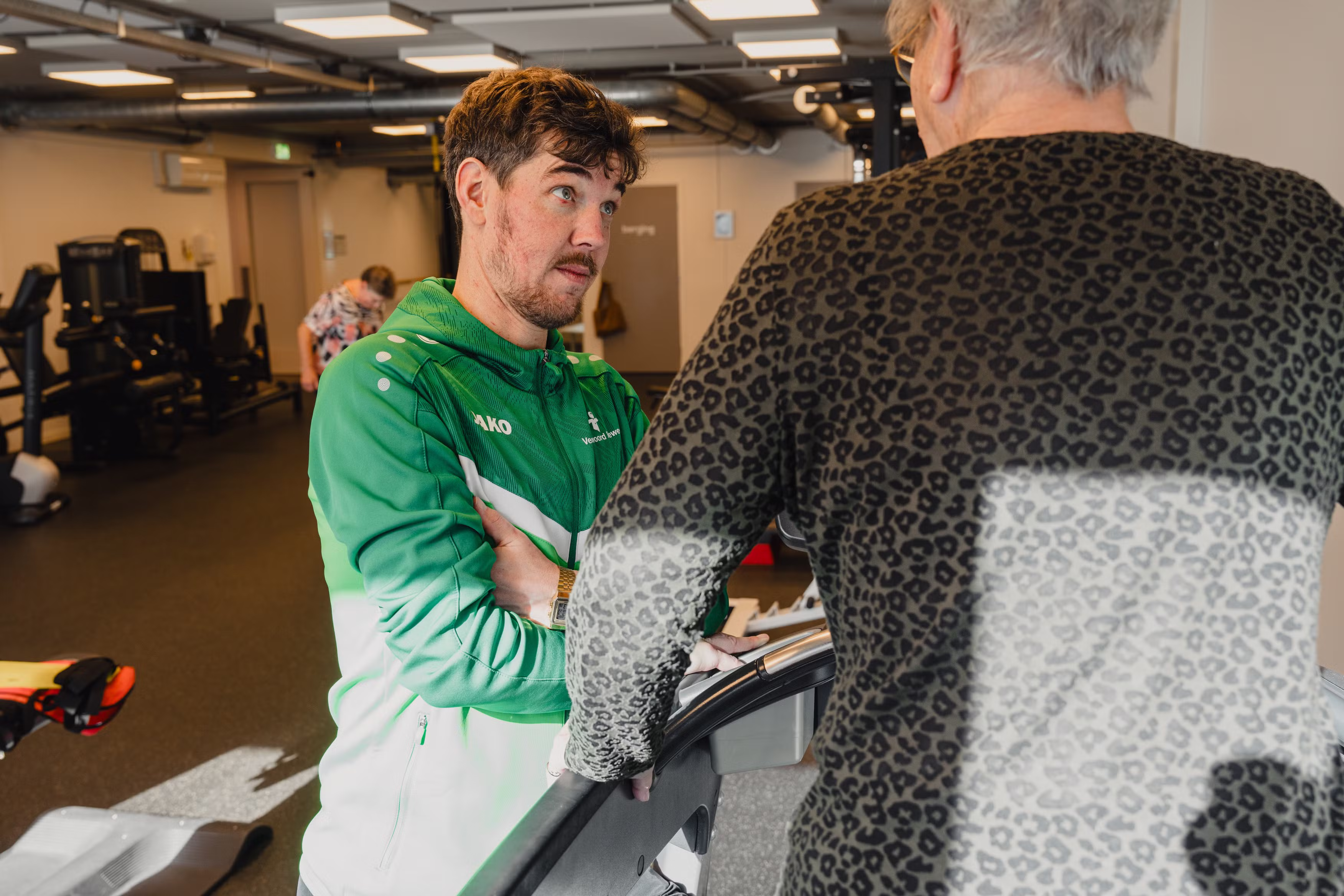 Young man in green sports jacket attentively talking to an older person wearing a leopard print top in a gym.