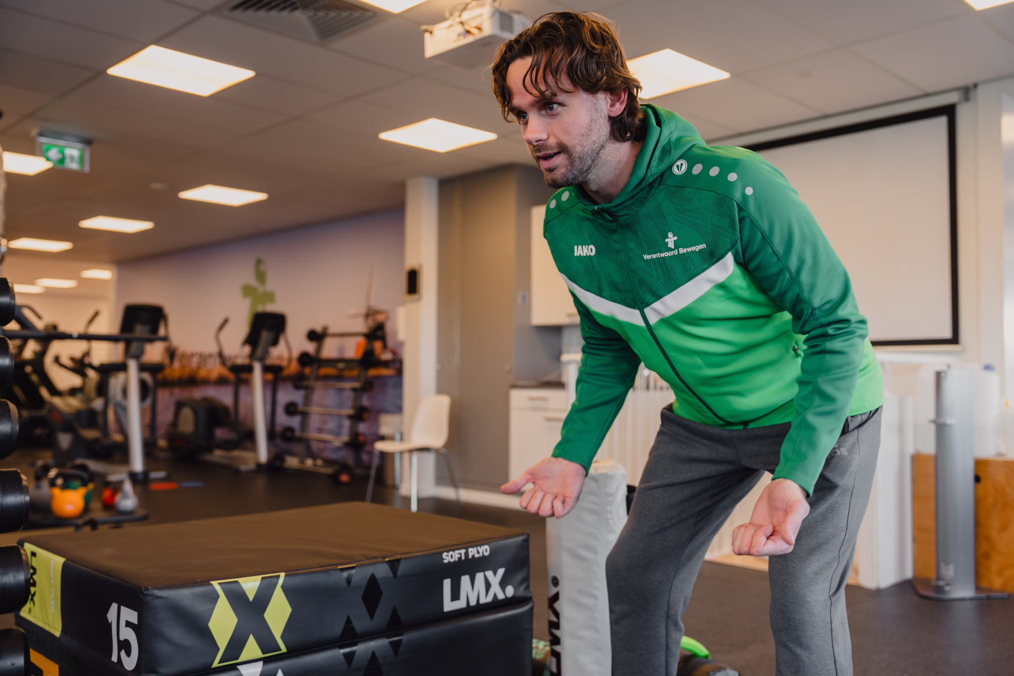 Man in green sports jacket and gray sweatpants standing in a gym next to a soft plyo box labeled 15 LMX.