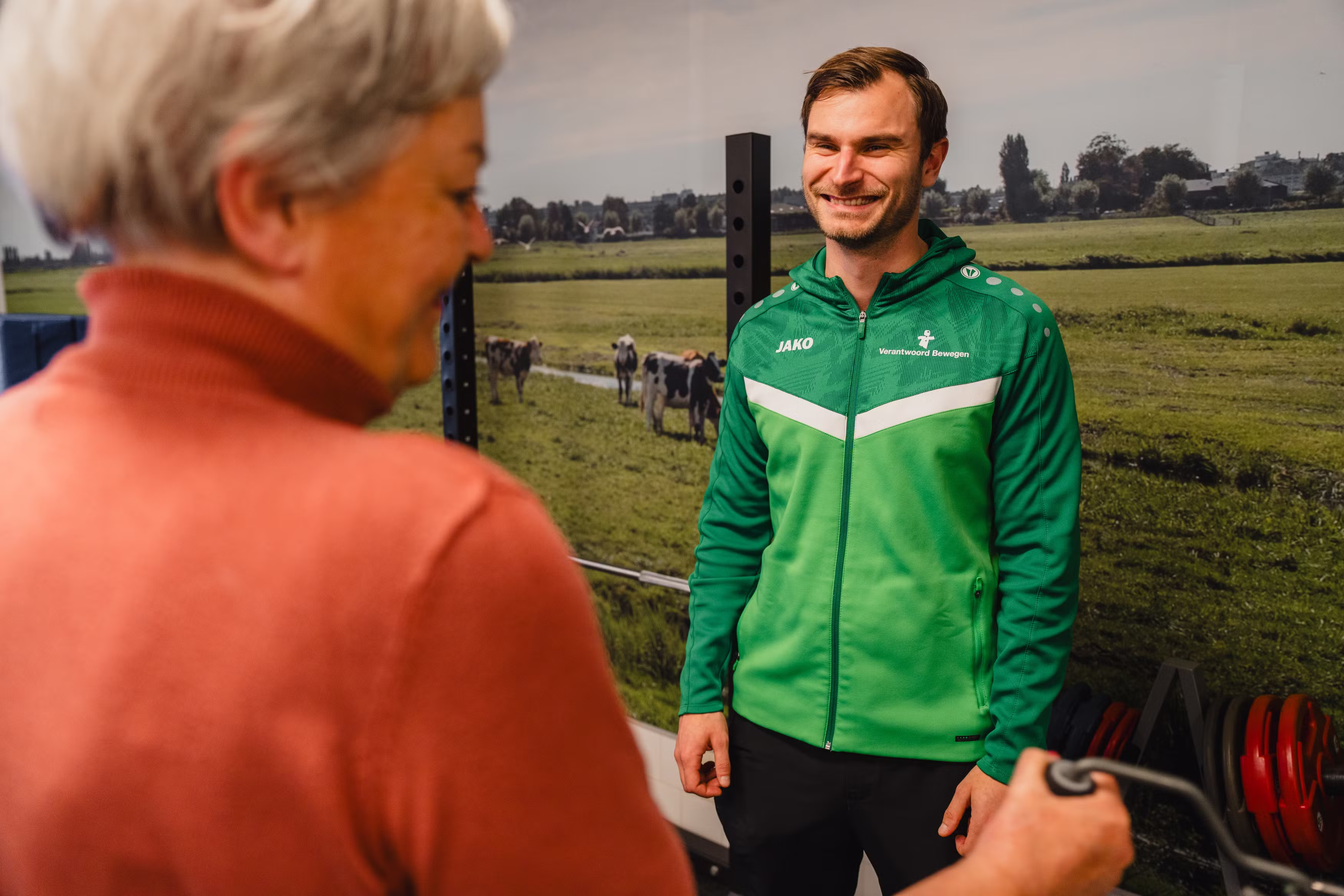 Smiling man in a green sports jacket standing indoors near a woman in a red sweater holding gym equipment, with a mural of cows in a green field in the background.