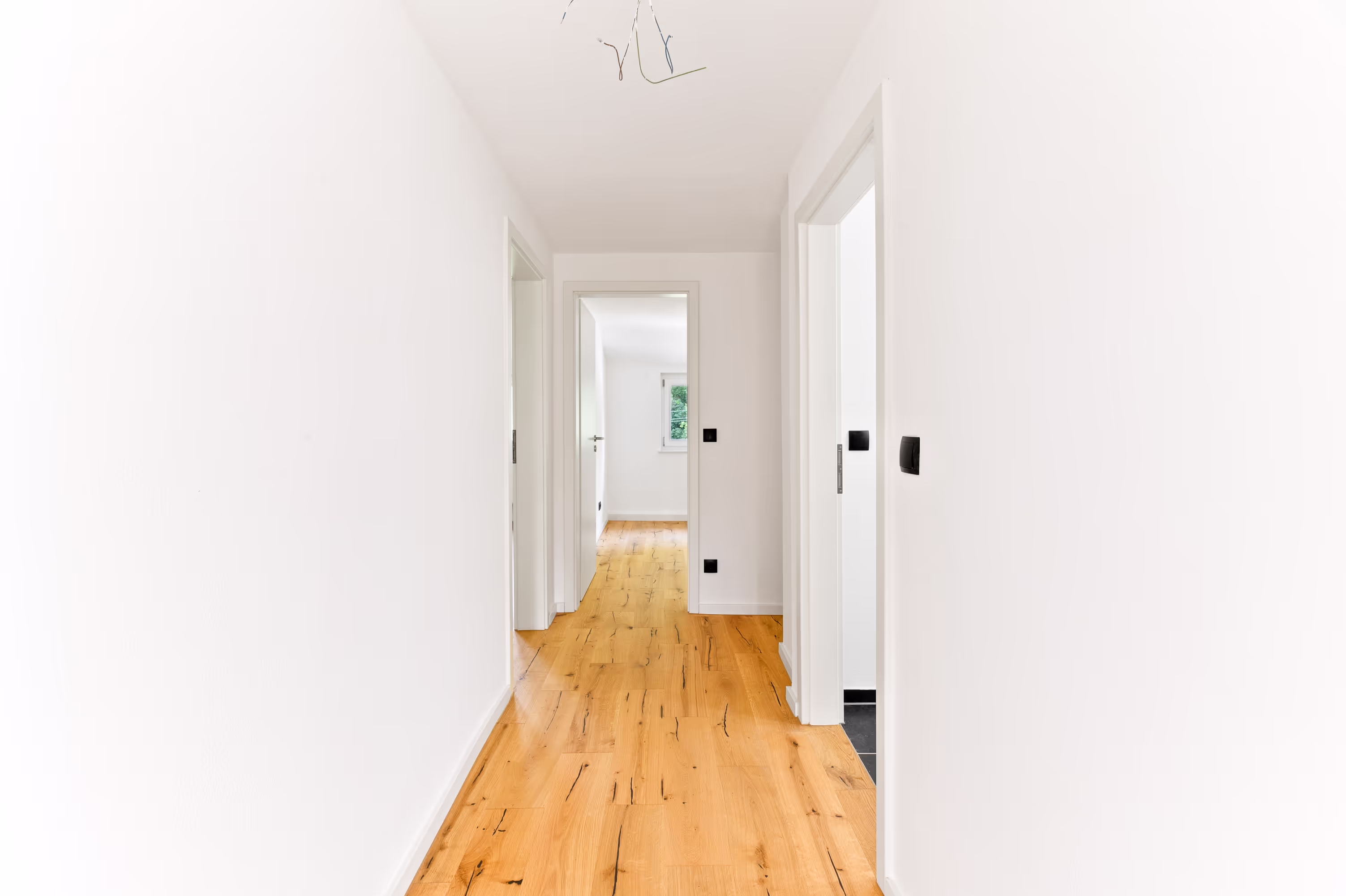 Bright hallway with natural wooden flooring, white walls, and multiple doors leading to rooms in a modern apartment.