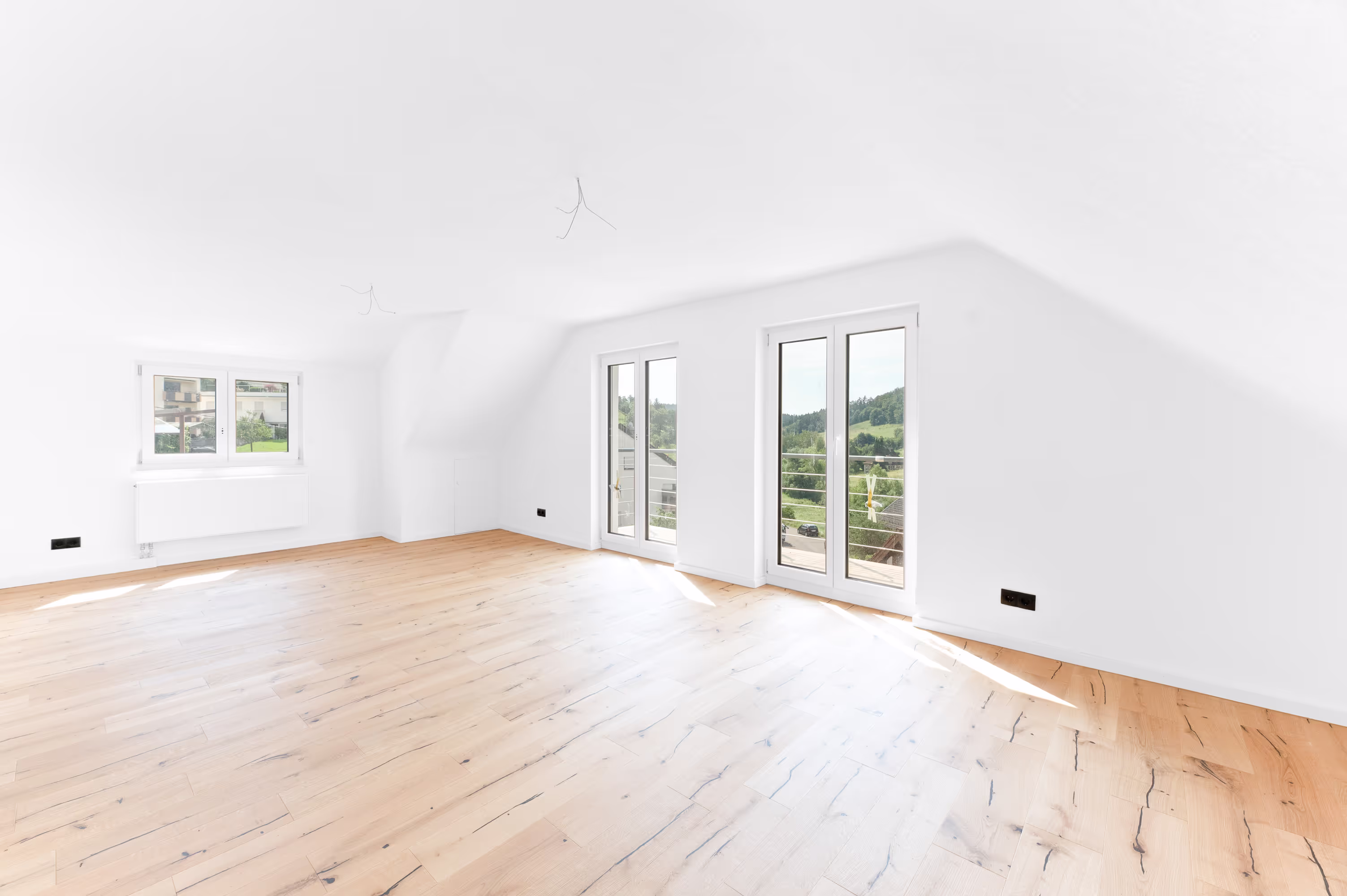 Spacious, sunlit living room with wooden flooring, large windows, and a scenic view of greenery outside.