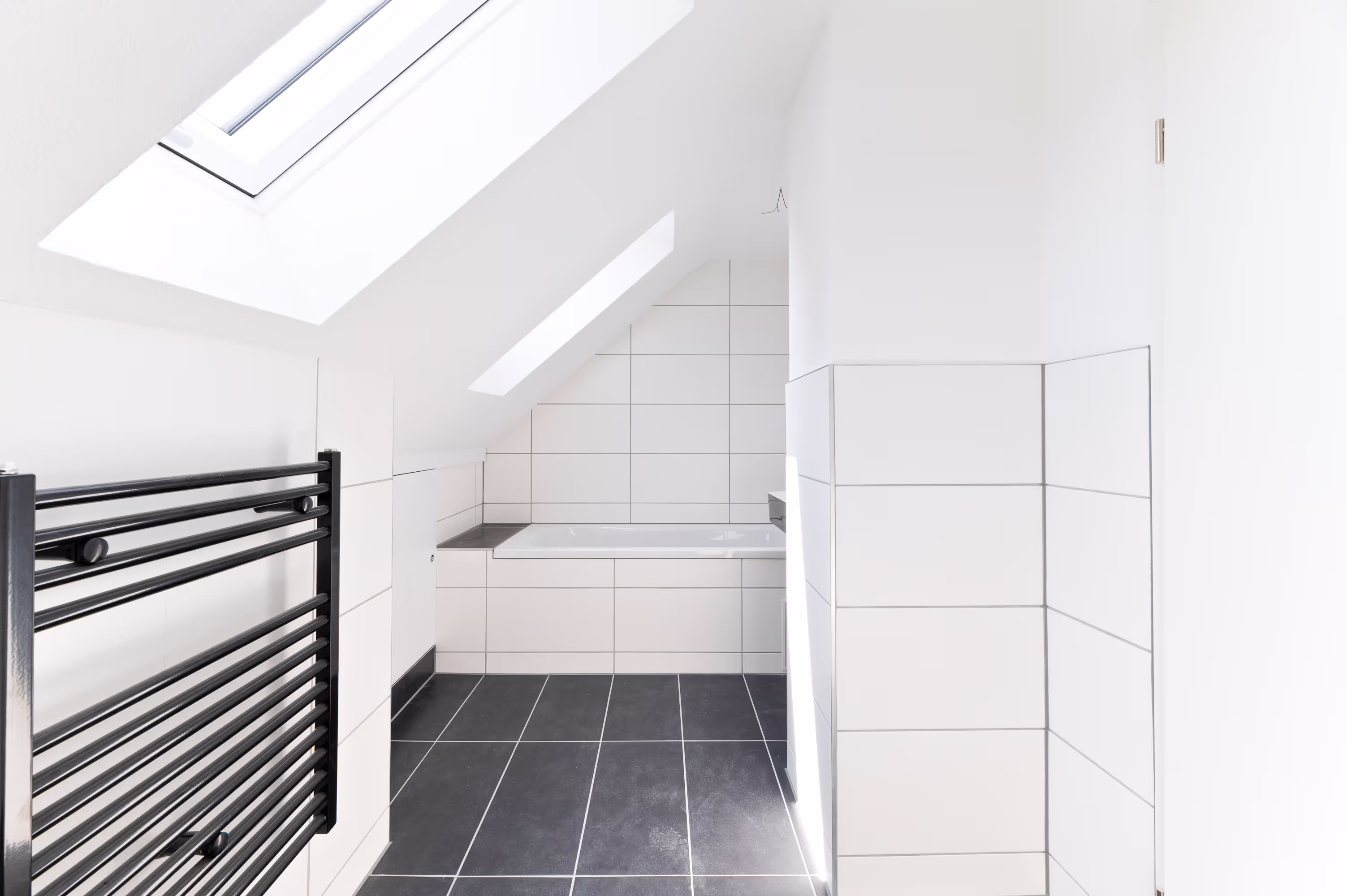 Modern bathroom with black towel radiator, skylights, white tiled walls, and a built-in bathtub under sloped ceilings.