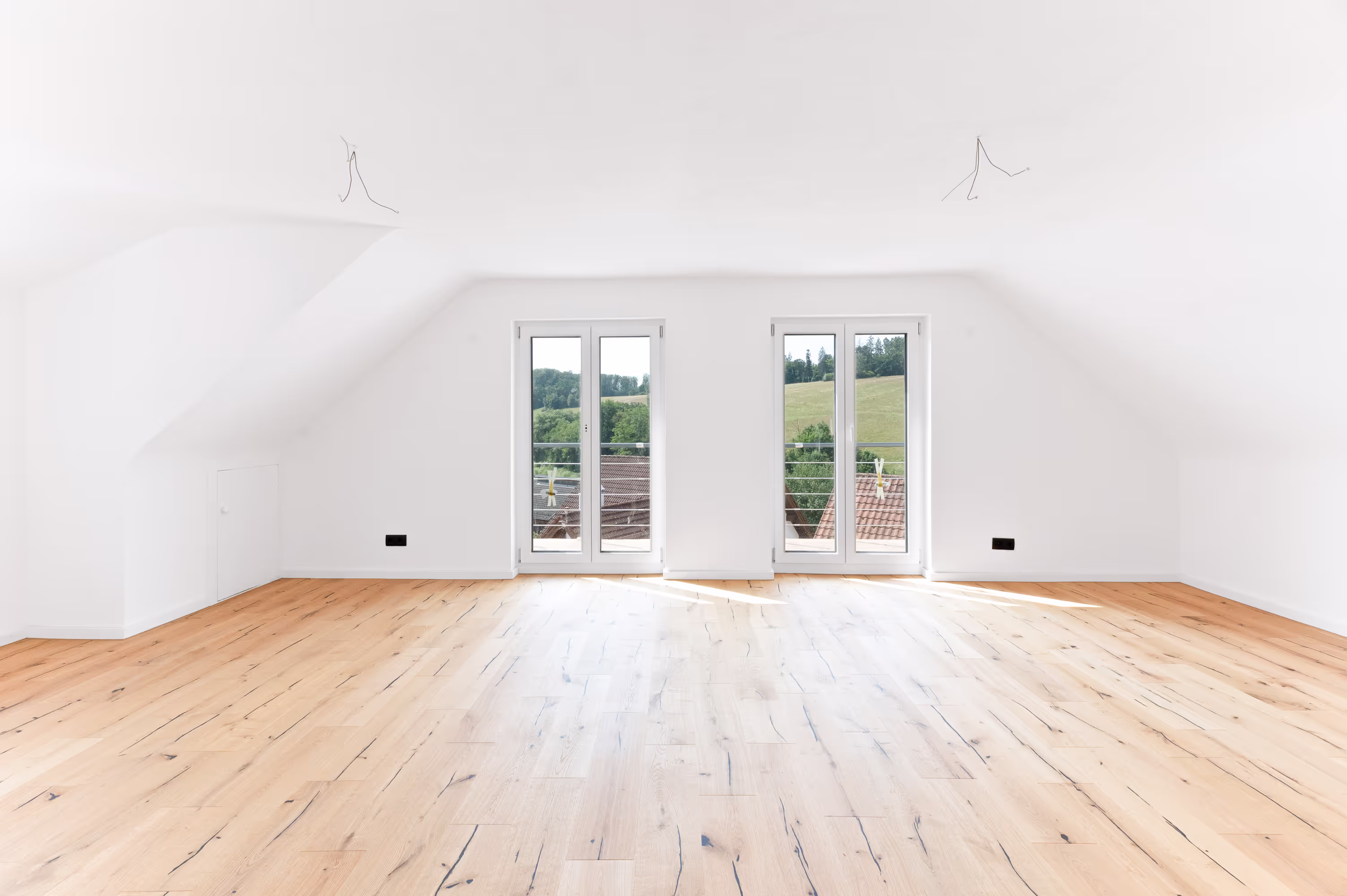 Bright and spacious living room with wooden flooring, two large balcony doors, and a scenic view of green hills and rooftops.